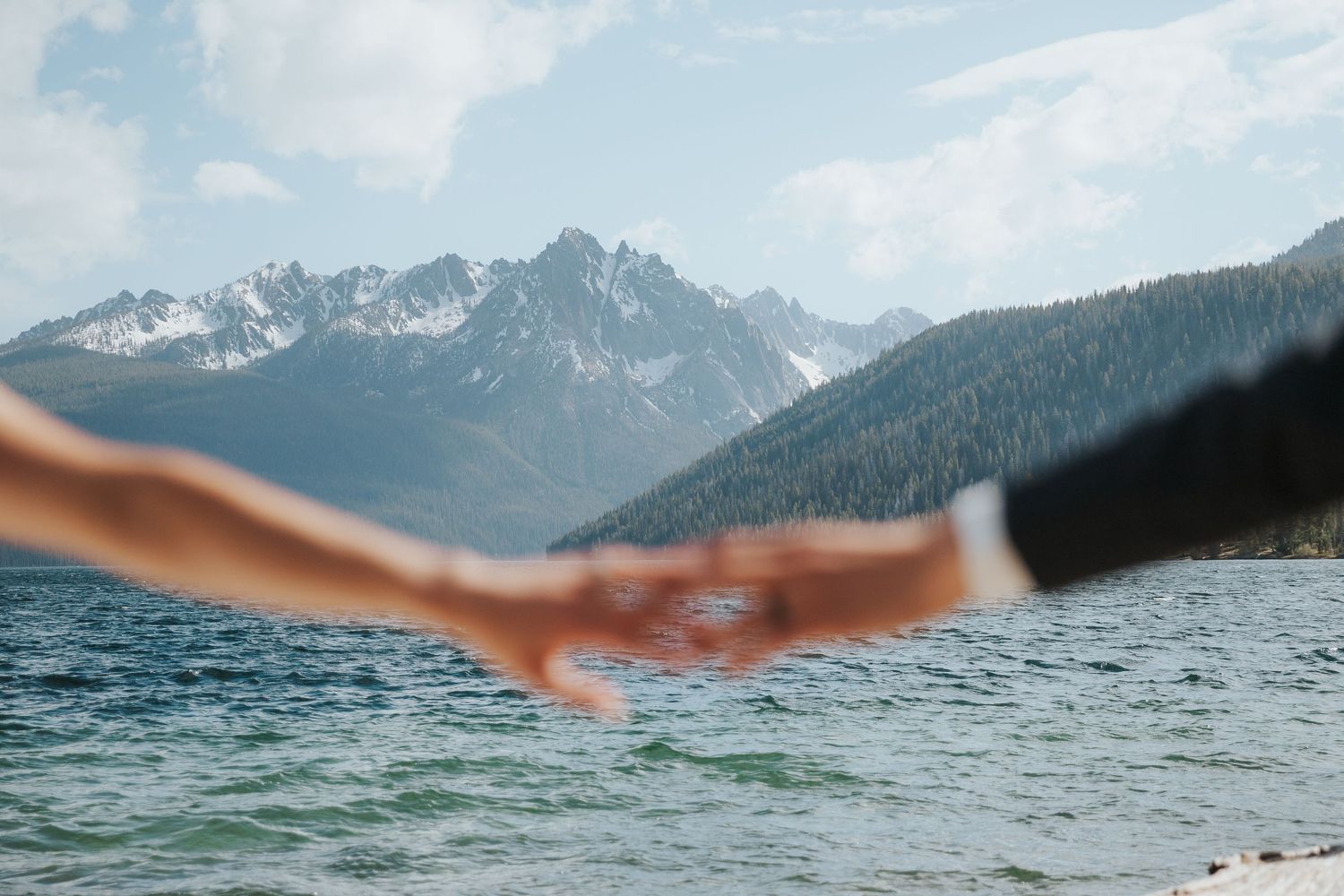 Hands reaching out across mountain lake with snow-capped peaks in background.