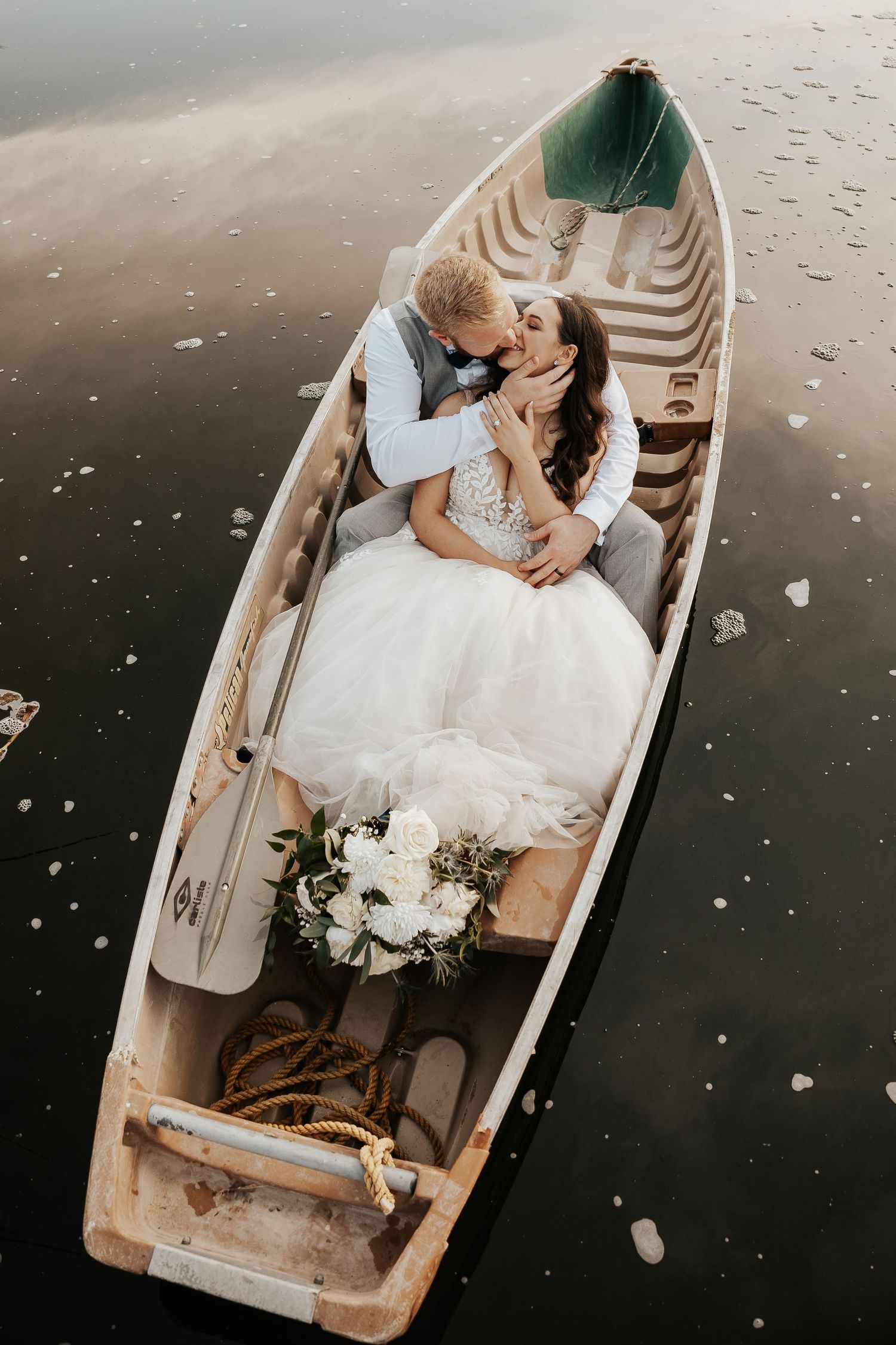 A romantic moment captured in a wooden rowboat on calm waters with wedding attire and floral details.