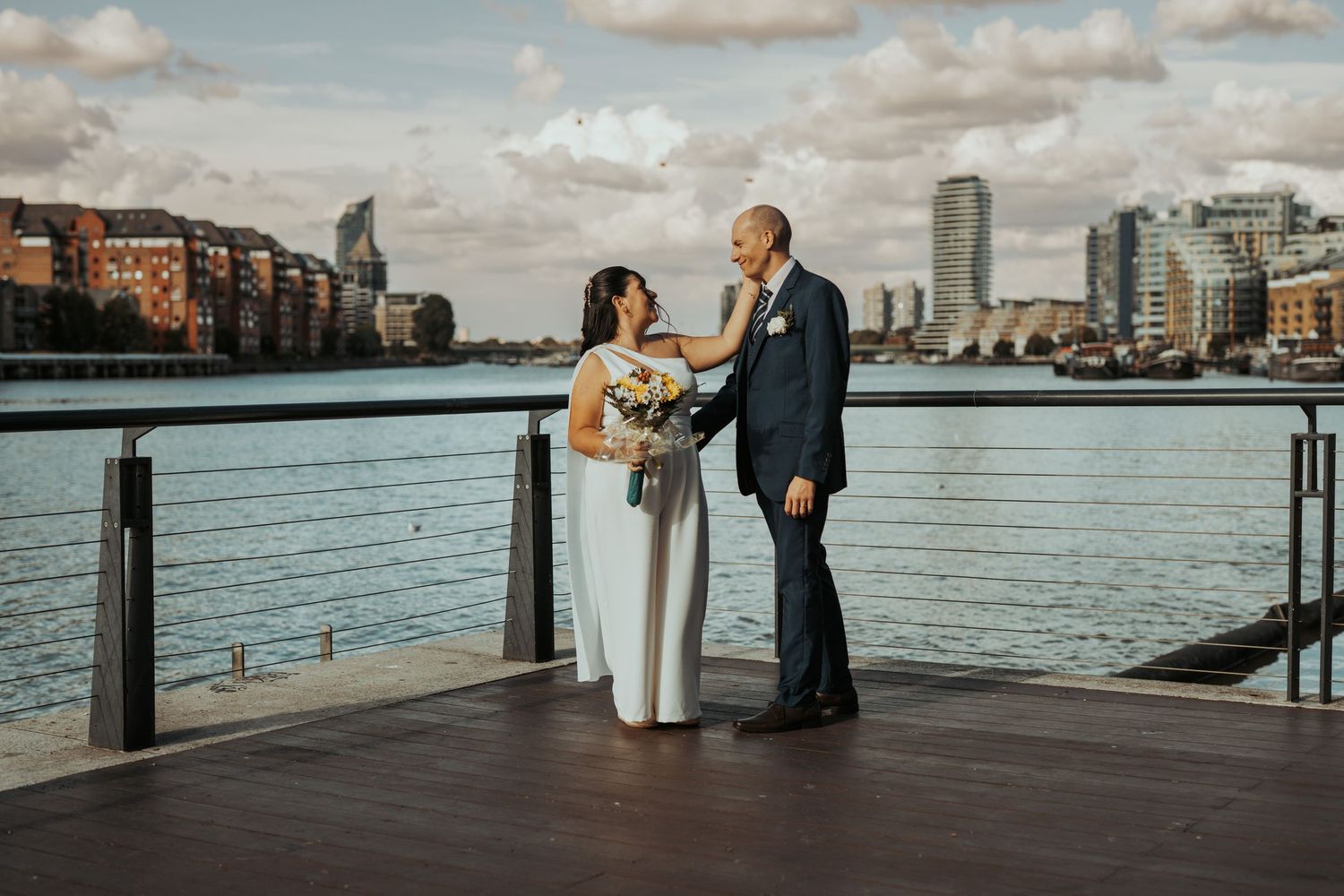 A couple stands together on a waterfront deck with the Boston skyline and harbor visible in the background.