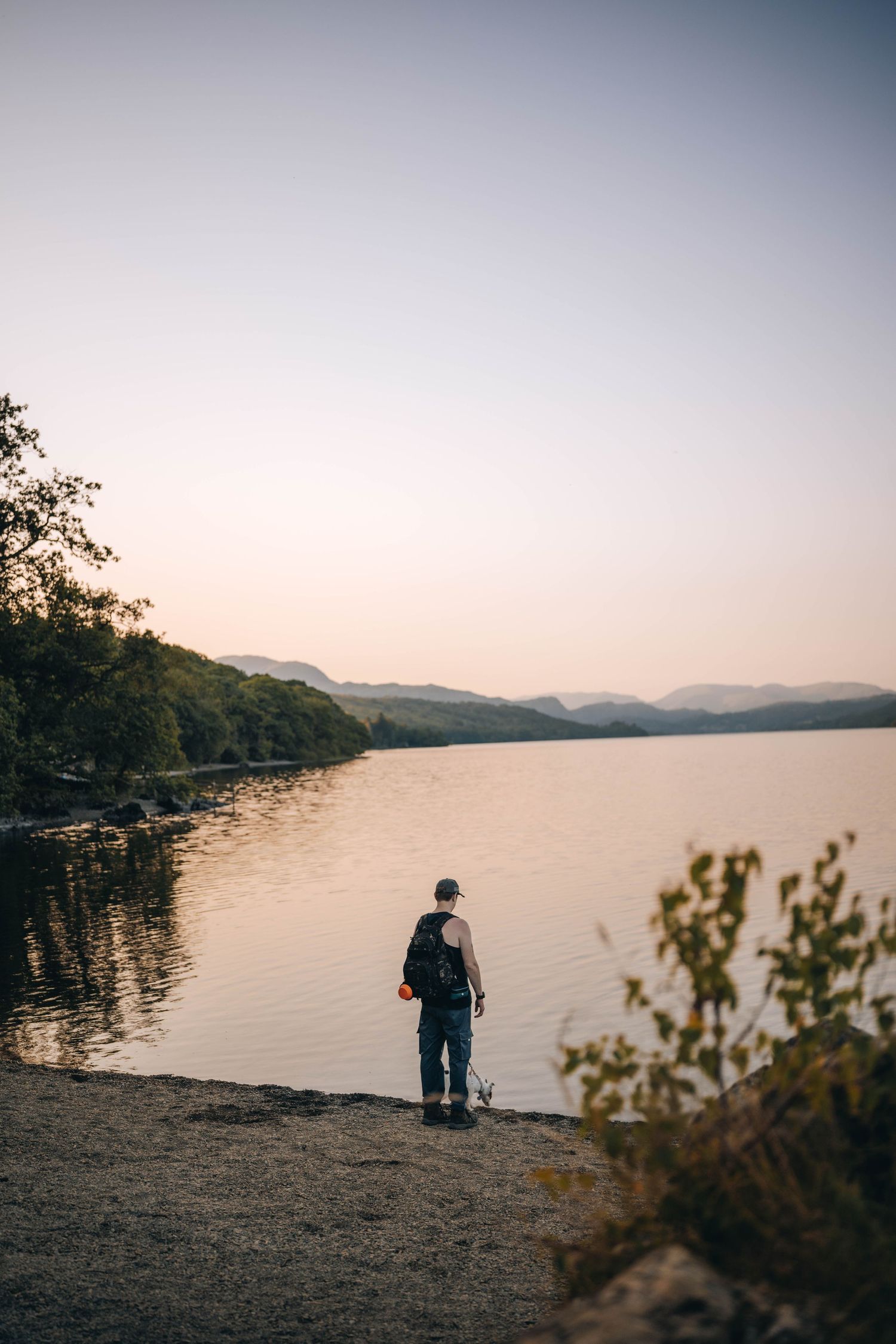 A lone hiker with a backpack stands on a rocky shore gazing at a calm lake surrounded by forested hills at sunset.