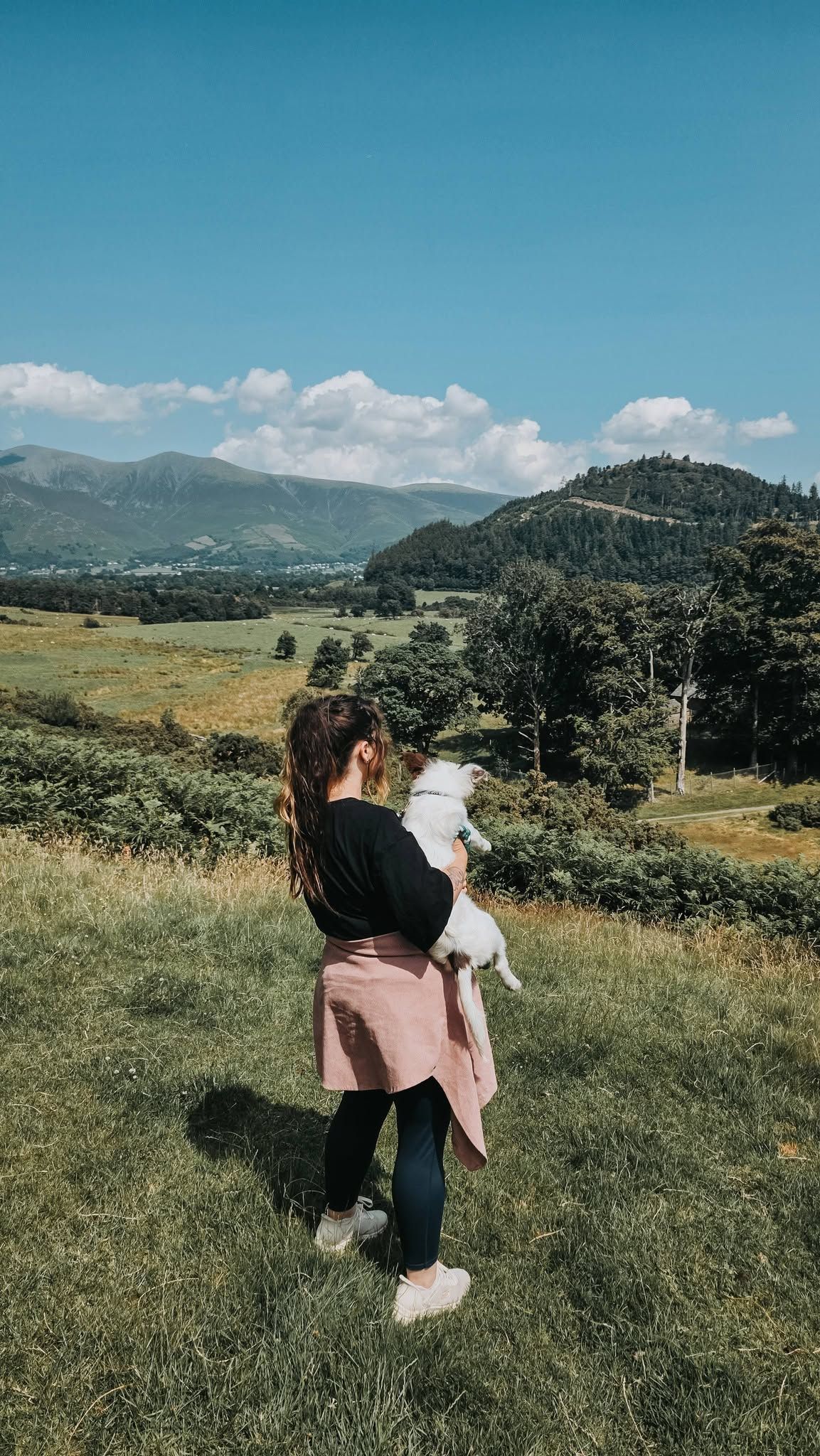 A woman holding a small white dog stands on a grassy hill with a scenic mountain valley and blue sky behind her.