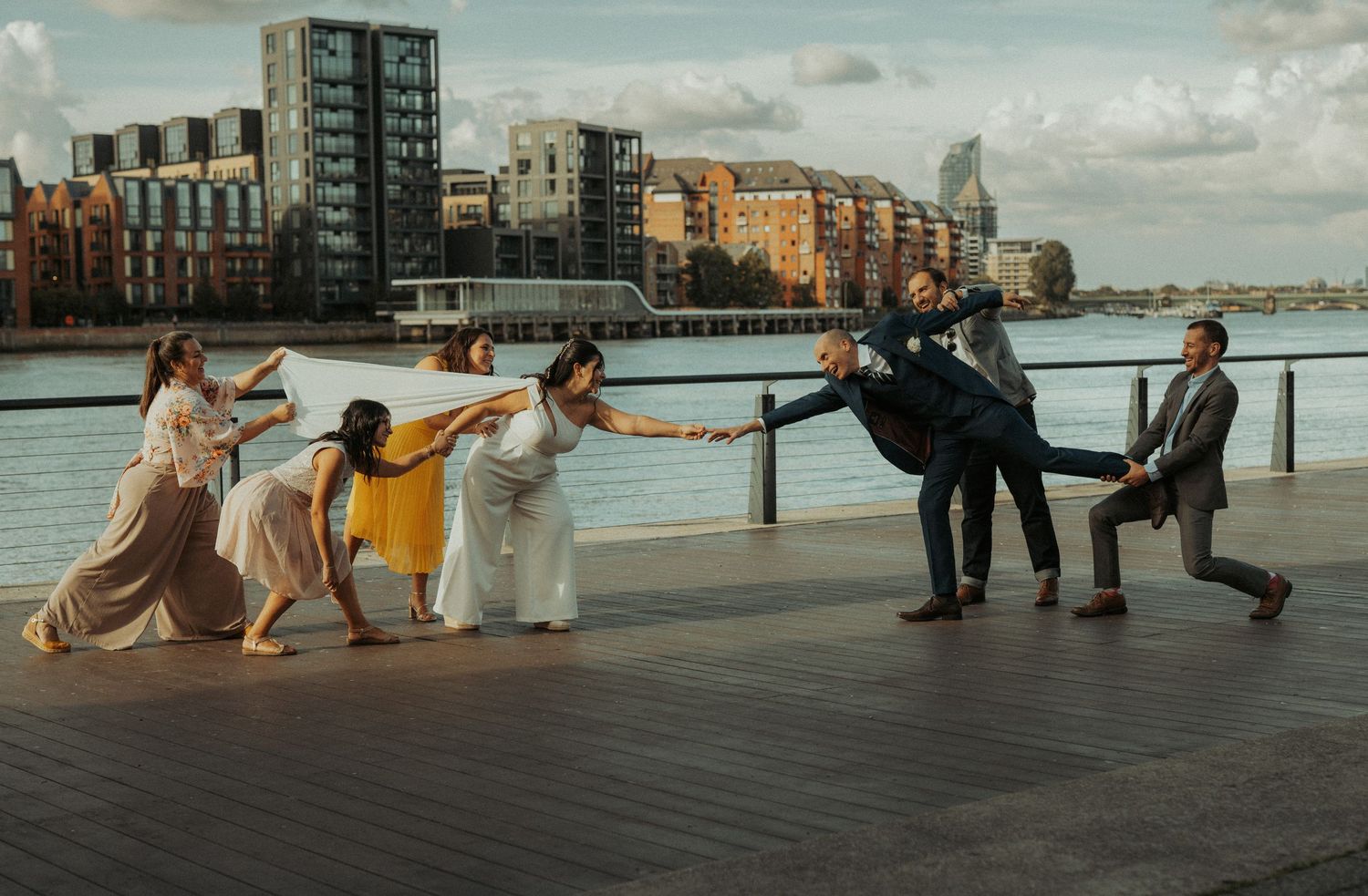 A group of dancers perform a synchronized movement sequence along a waterfront with urban buildings in the background.