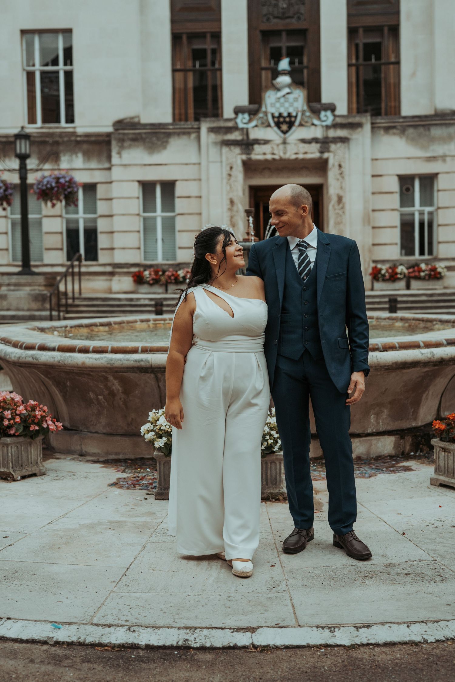 A couple in wedding attire stands in front of an elegant historic building with ornate architecture and a fountain.