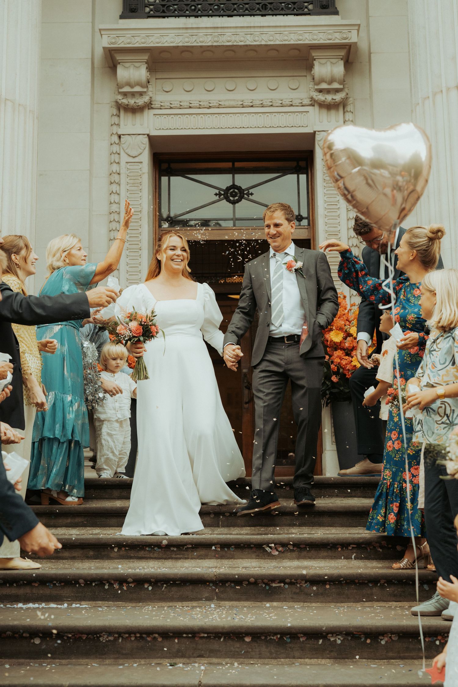 Wedding party gathered on stone steps outside venue with guests throwing confetti in celebration.