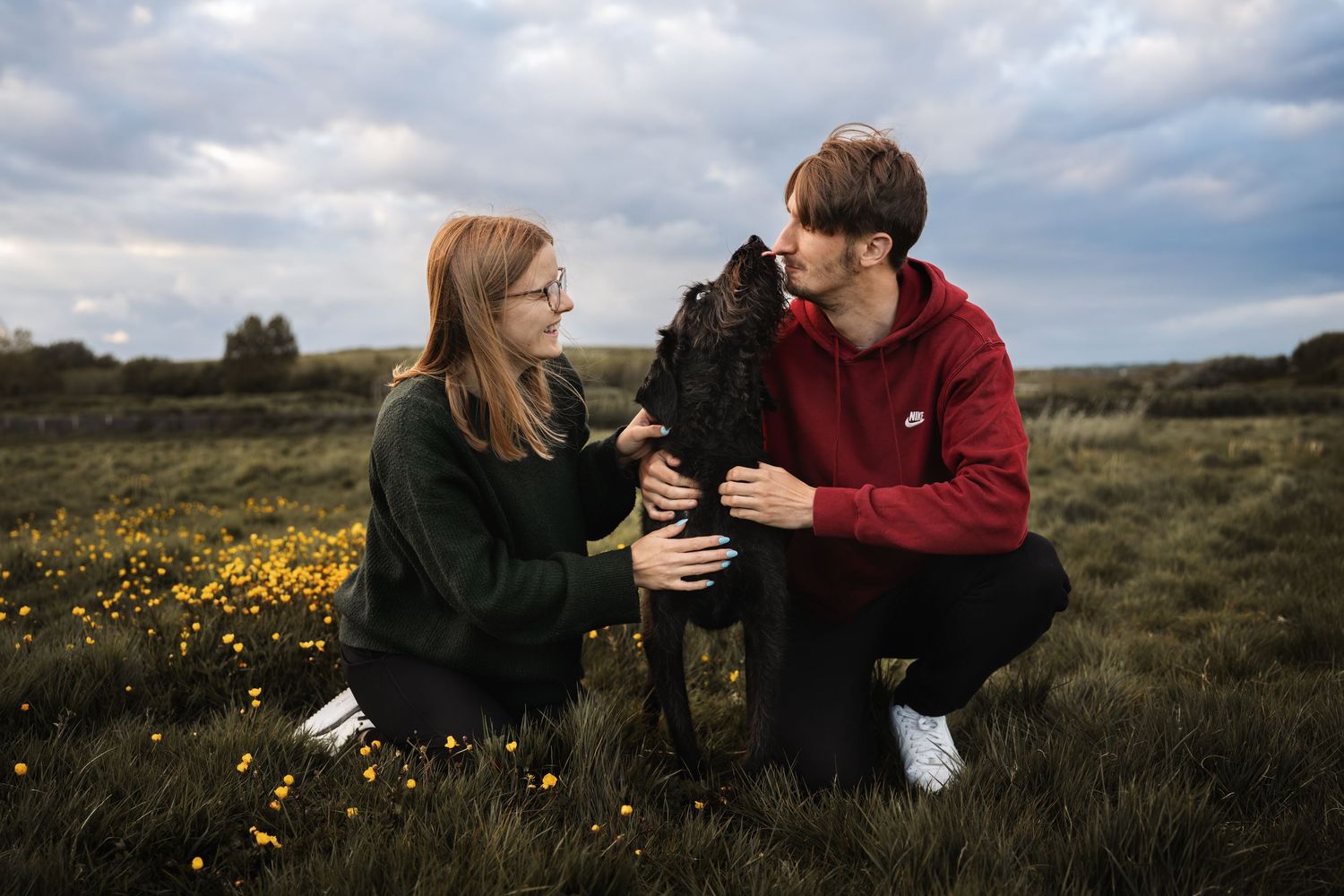 A couple crouching in a flower field petting their black curly-haired dog under a cloudy sky.
