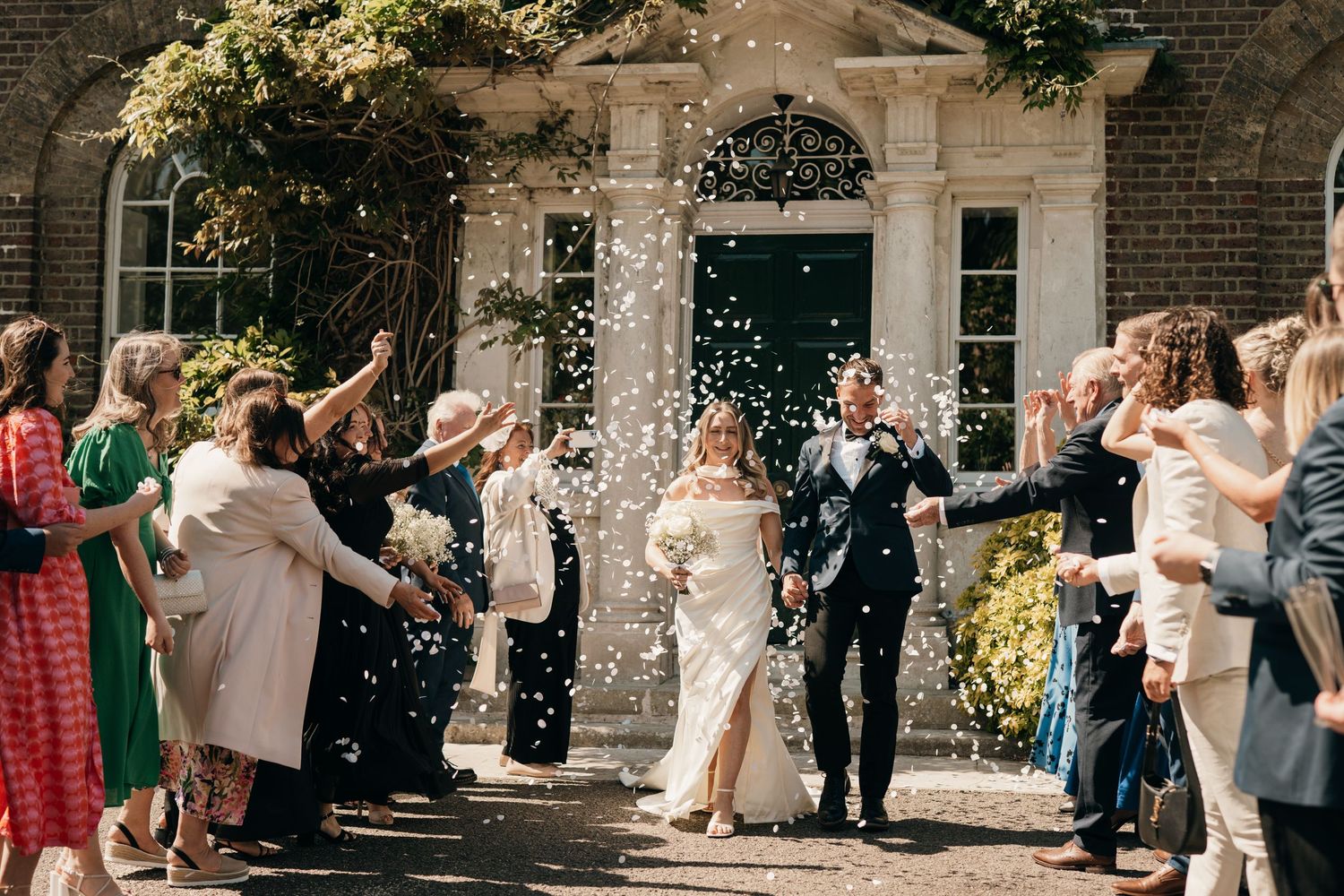 Wedding guests shower a couple with confetti as they exit a historic brick building with stone archway.