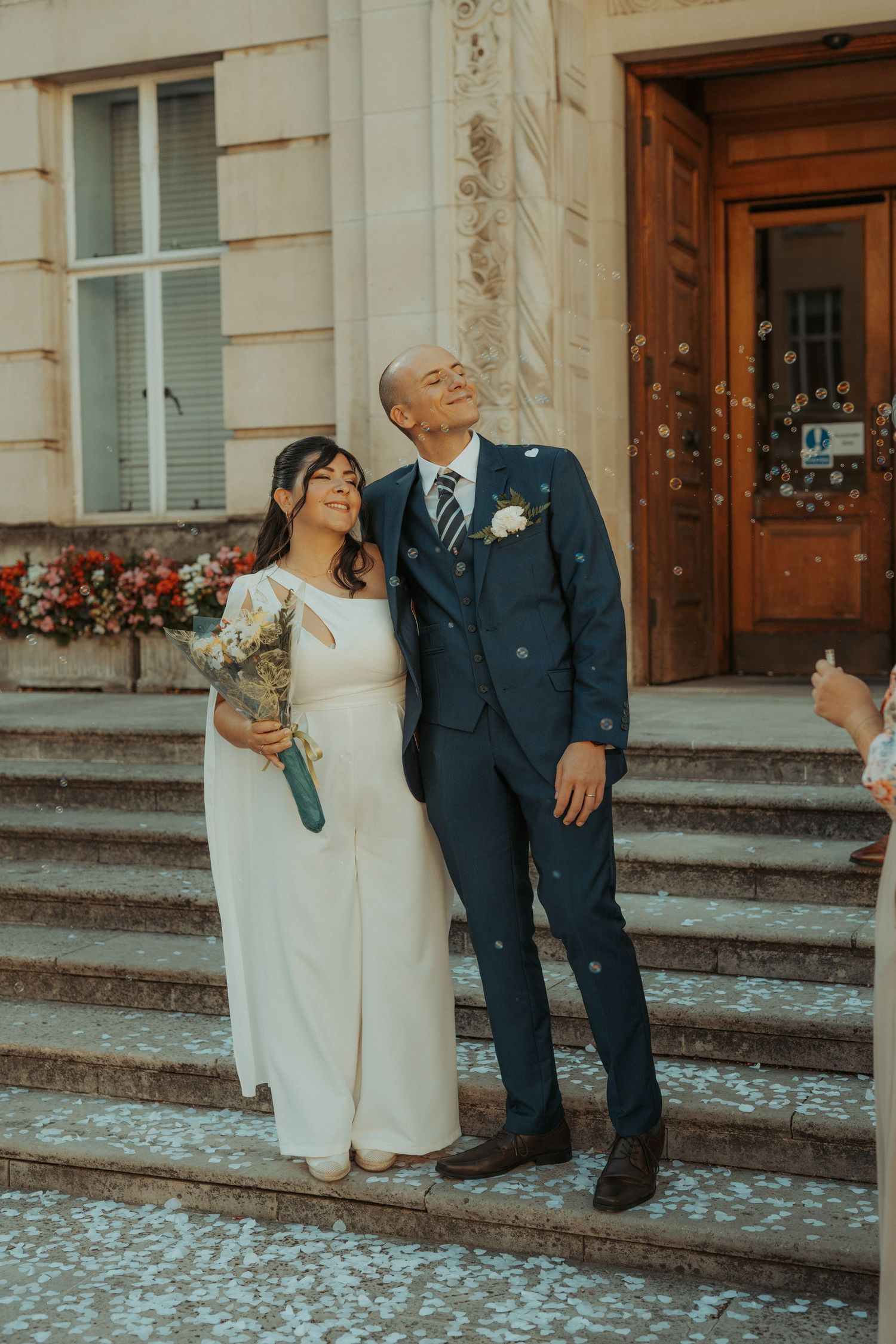 A newlywed couple shares a joyful moment on stone steps outside a building with a wooden door and floral decorations.