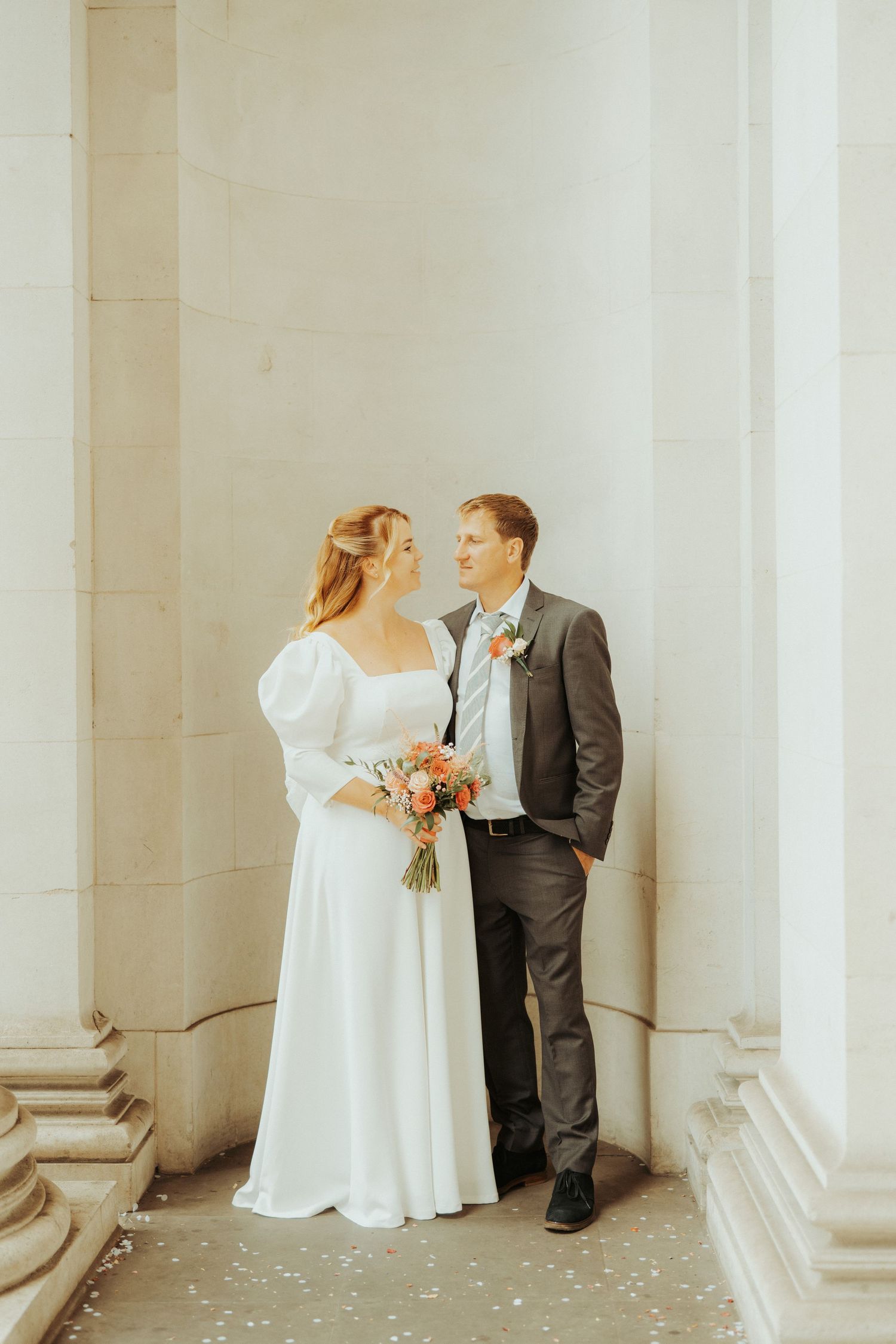 A couple in wedding attire stands between elegant white columns, with the bride holding a peach-colored bouquet.
