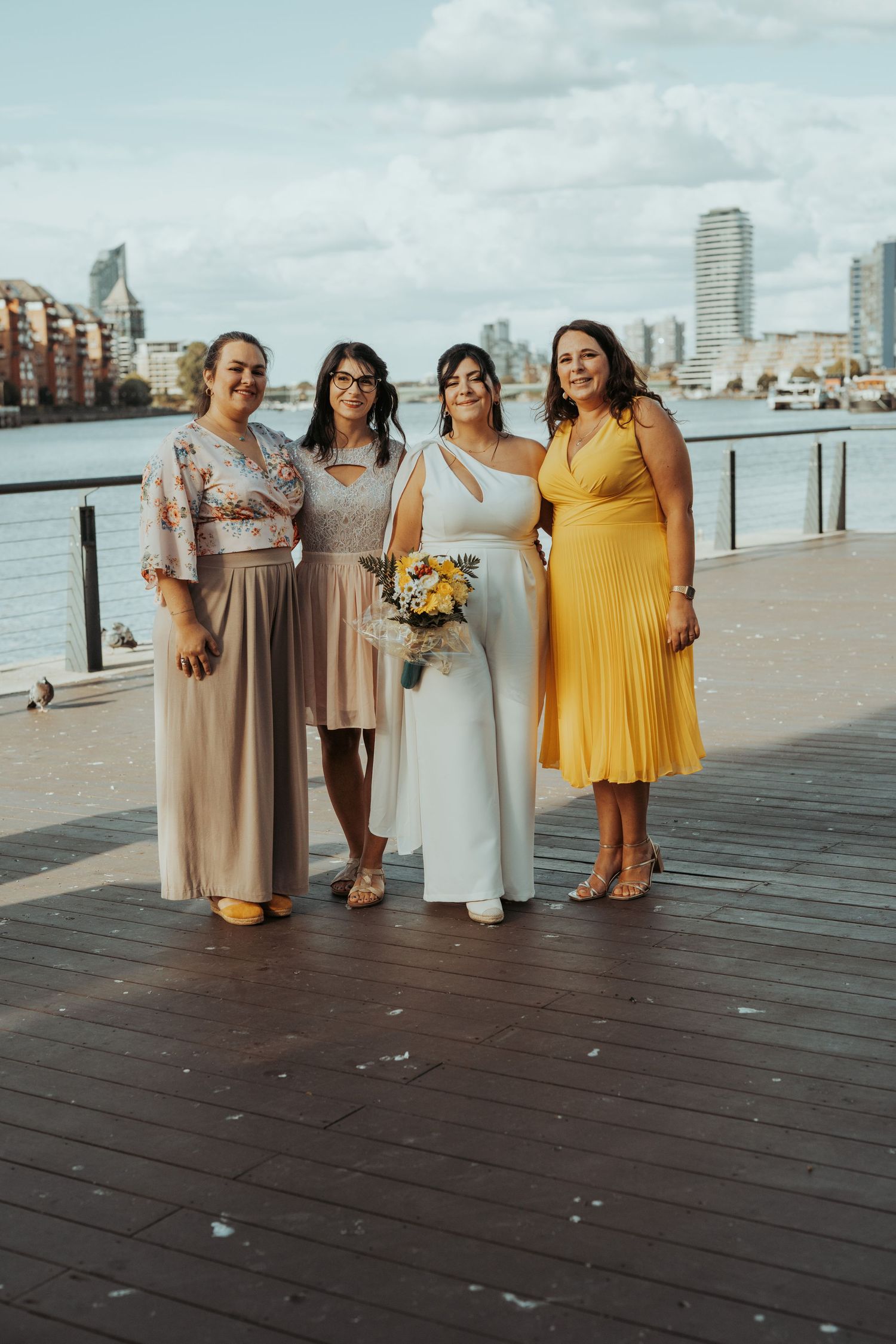 Wedding party in coordinated pastel dresses poses on waterfront boardwalk with city skyline in background.