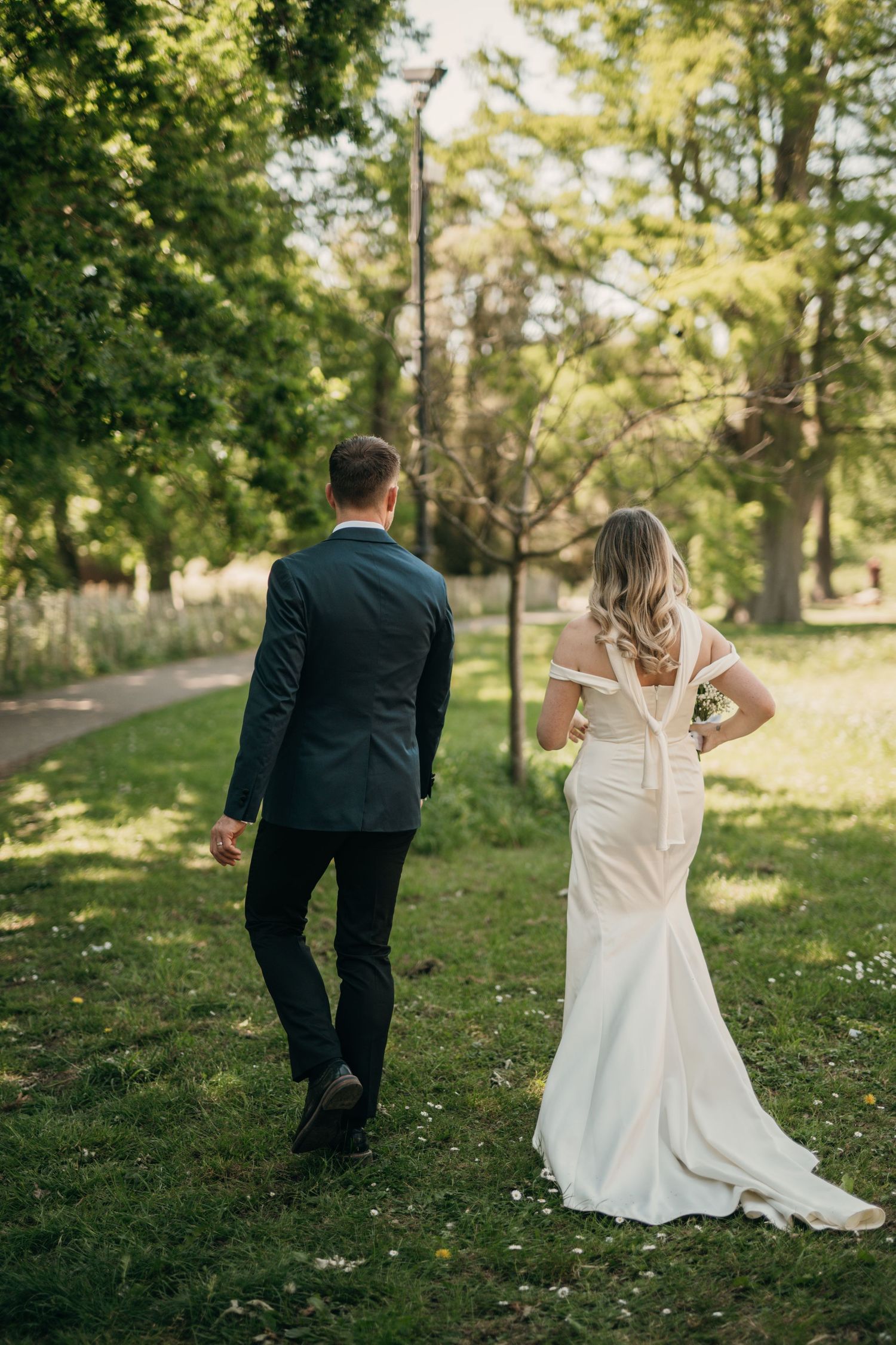 A couple in wedding attire walks through a sunlit wooded area during their outdoor wedding photoshoot.