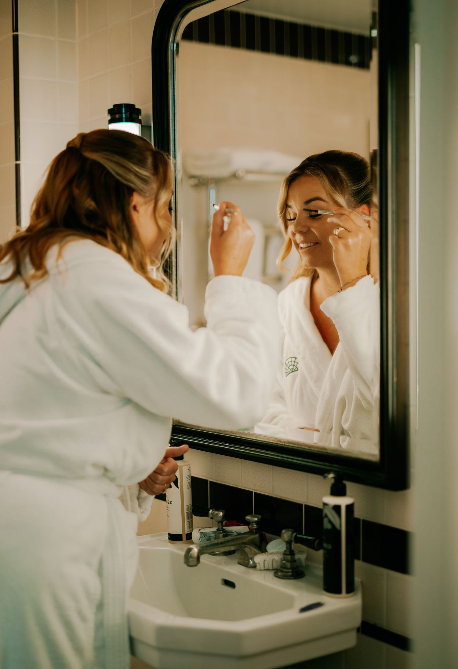 Someone in a white bathrobe applies makeup while looking in a bathroom mirror during morning preparations.