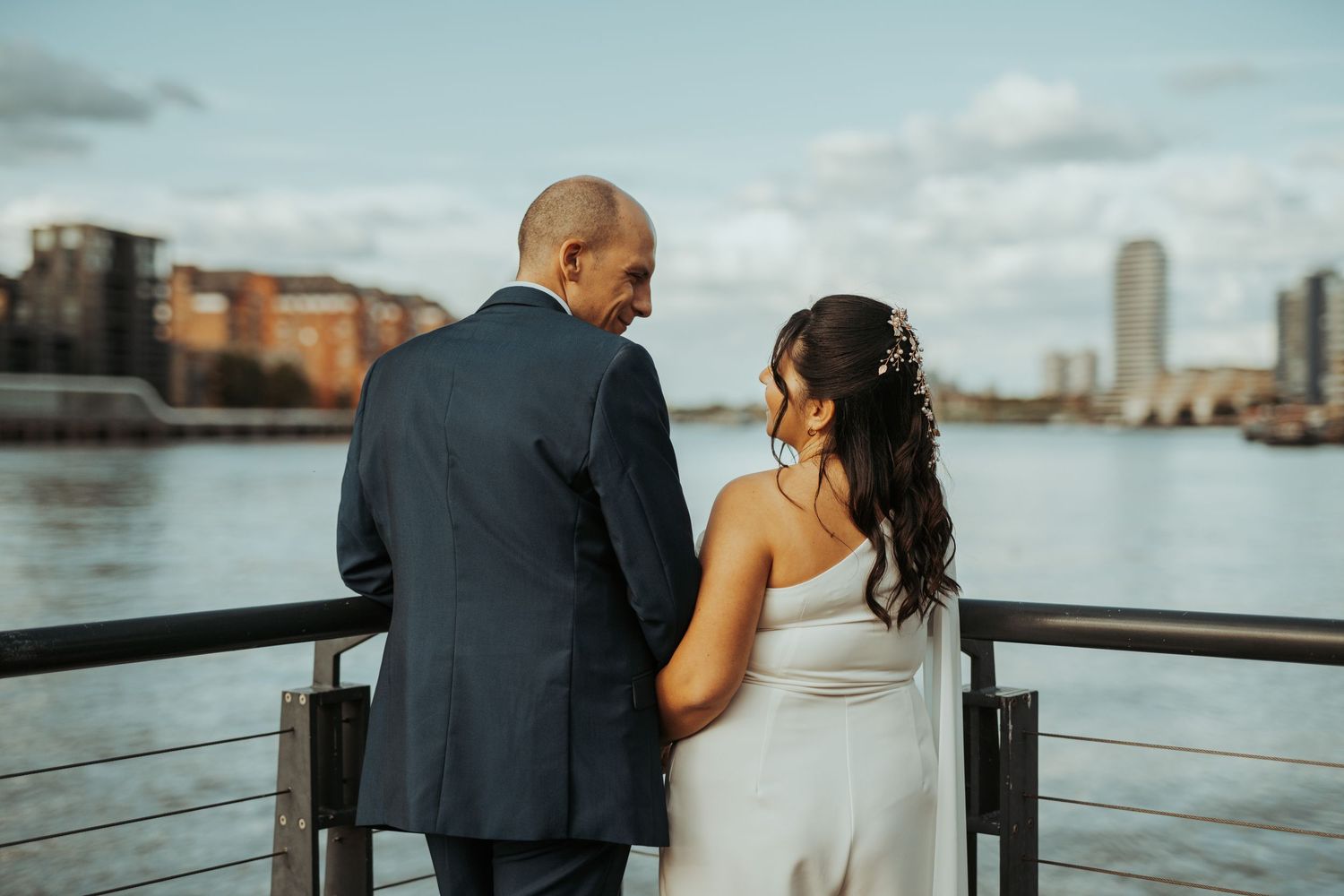 A couple shares a romantic moment on a waterfront deck overlooking a city harbor during their wedding day.
