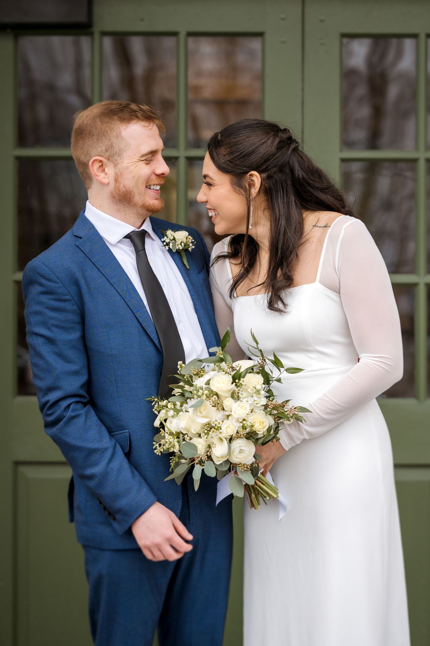 Wedding couple in blue suit and white dress posing with bouquet against green doors.