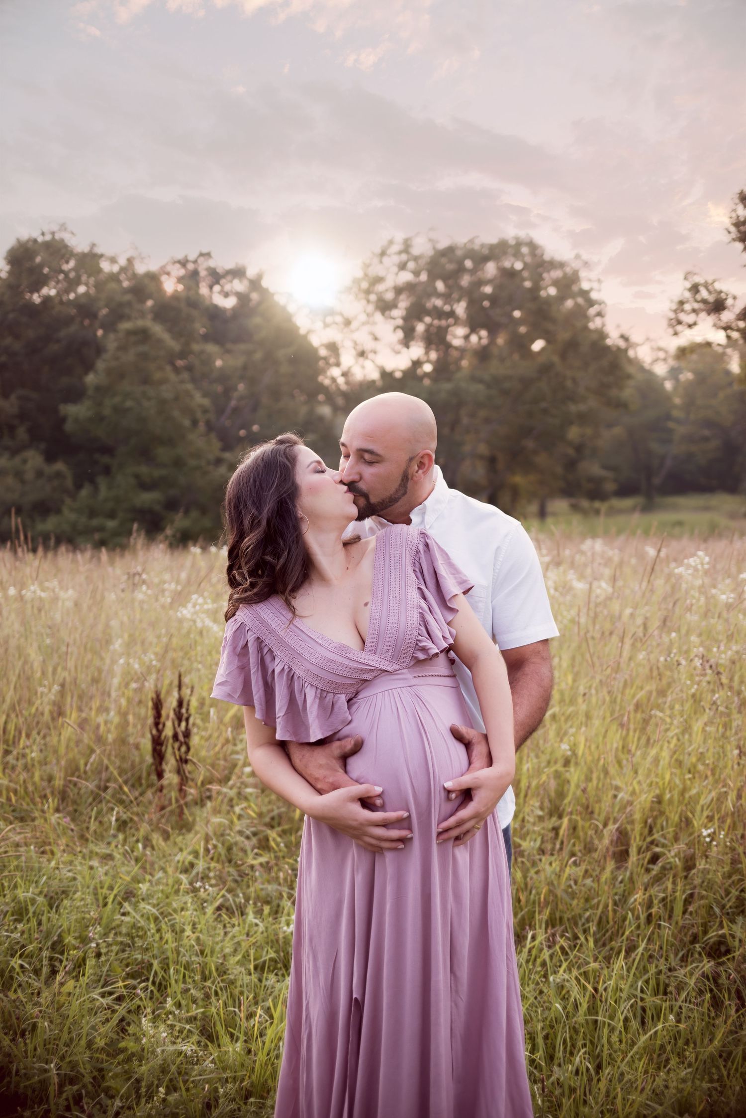 A couple shares an intimate moment in a field during sunset wearing coordinated mauve and white outfits.
