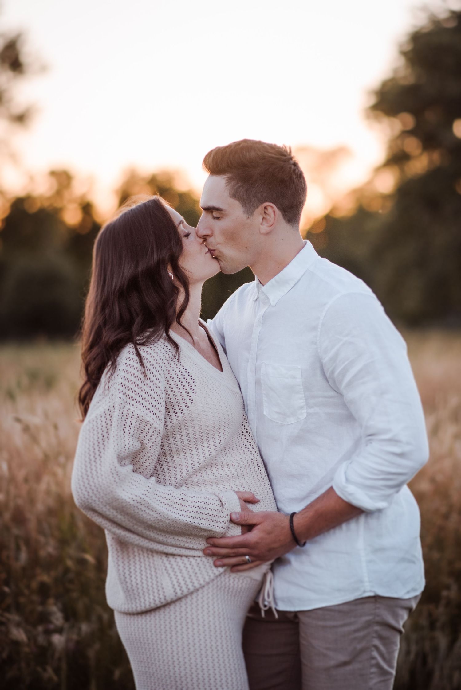 Romantic couple embracing in a field during sunset wearing white and neutral tones.