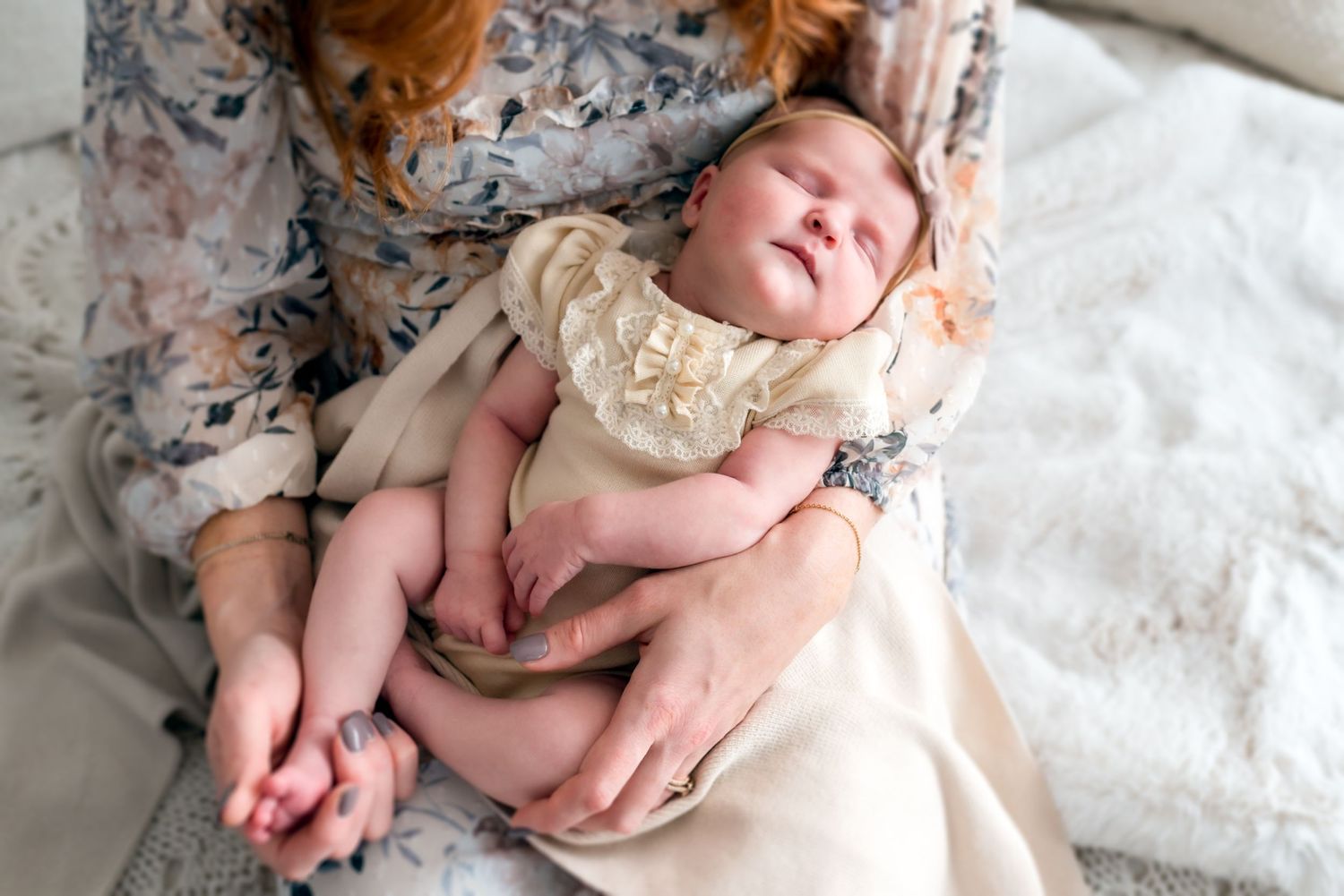Newborn baby wearing vintage-style lace dress resting on white bedding.