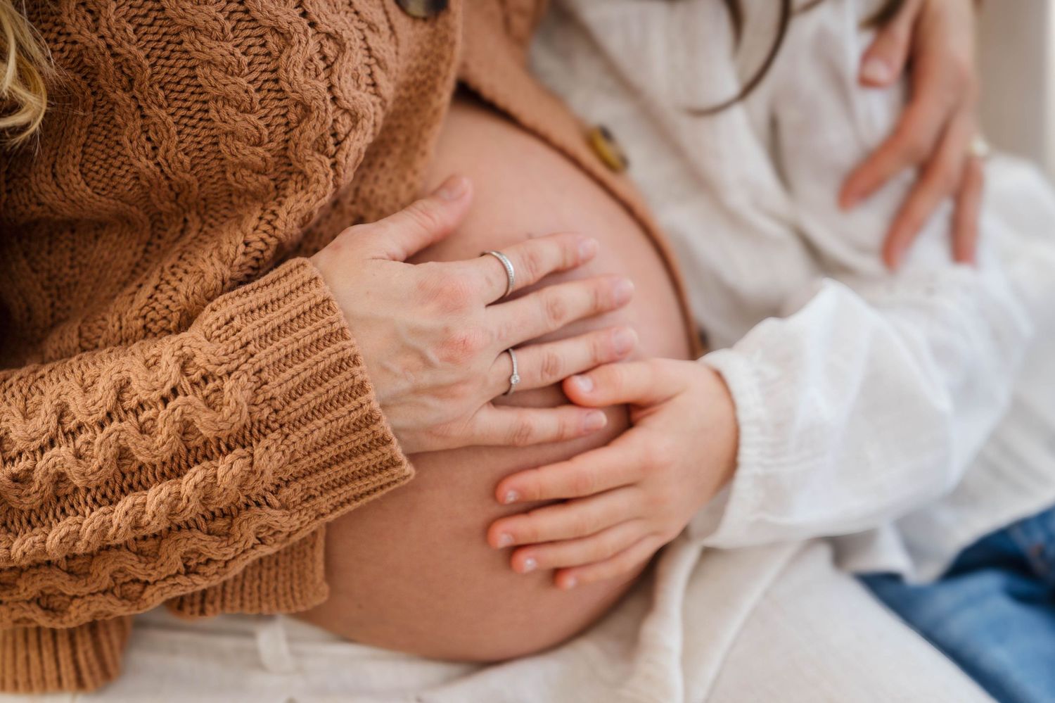 Close up of hands gently resting on a pregnant belly while wearing a brown knit sweater and white dress.