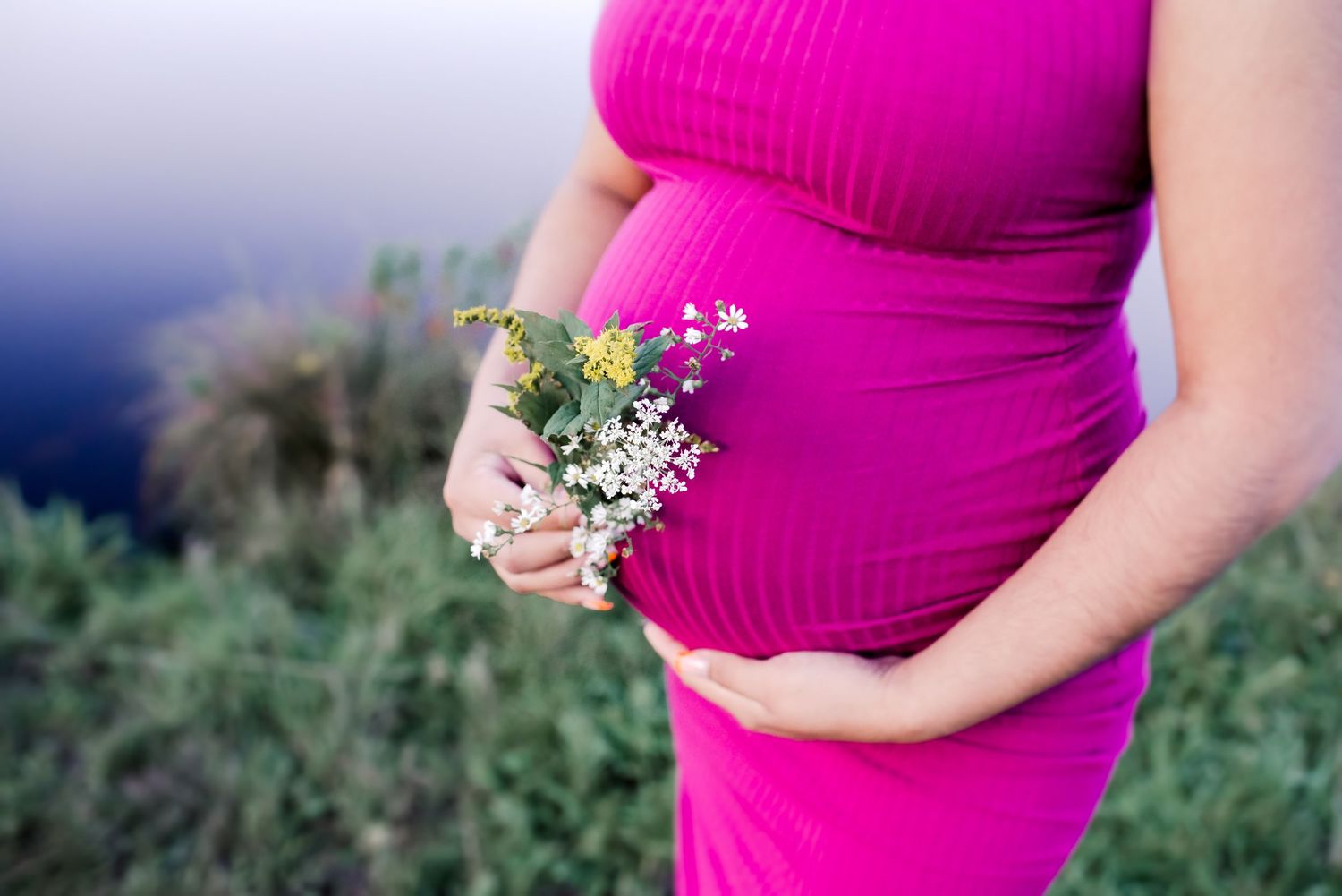 A person in a bright pink maternity dress poses against a natural outdoor background.