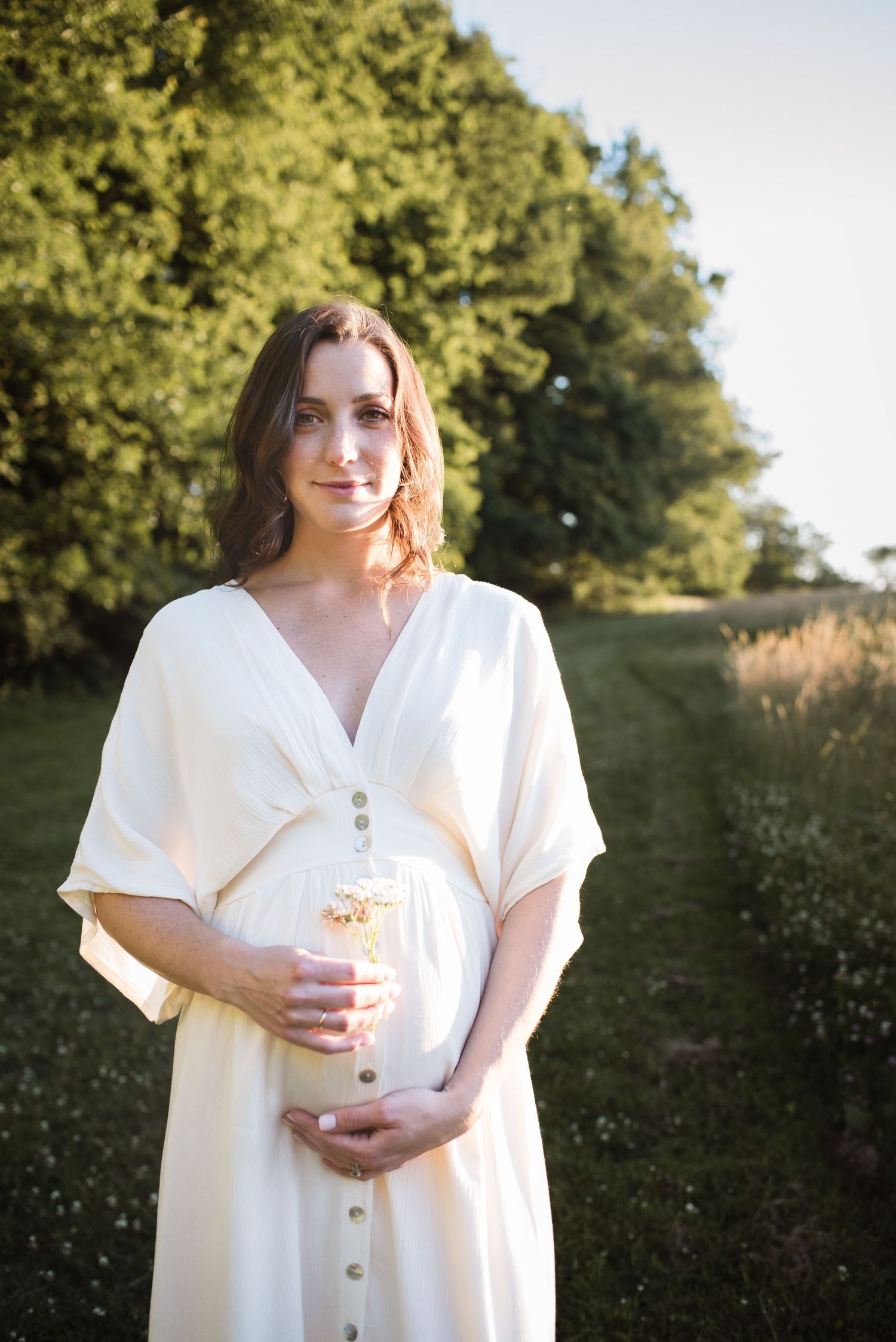 Woman in white dress holding drink while standing in sunlit field with wildflowers.