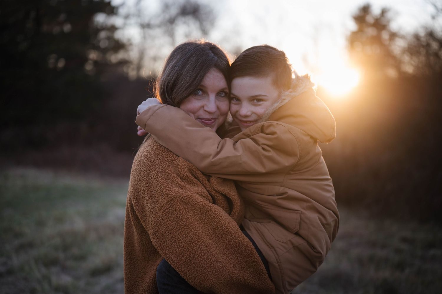 A tender embrace between two people wearing brown coats captured during golden hour sunset in a field.