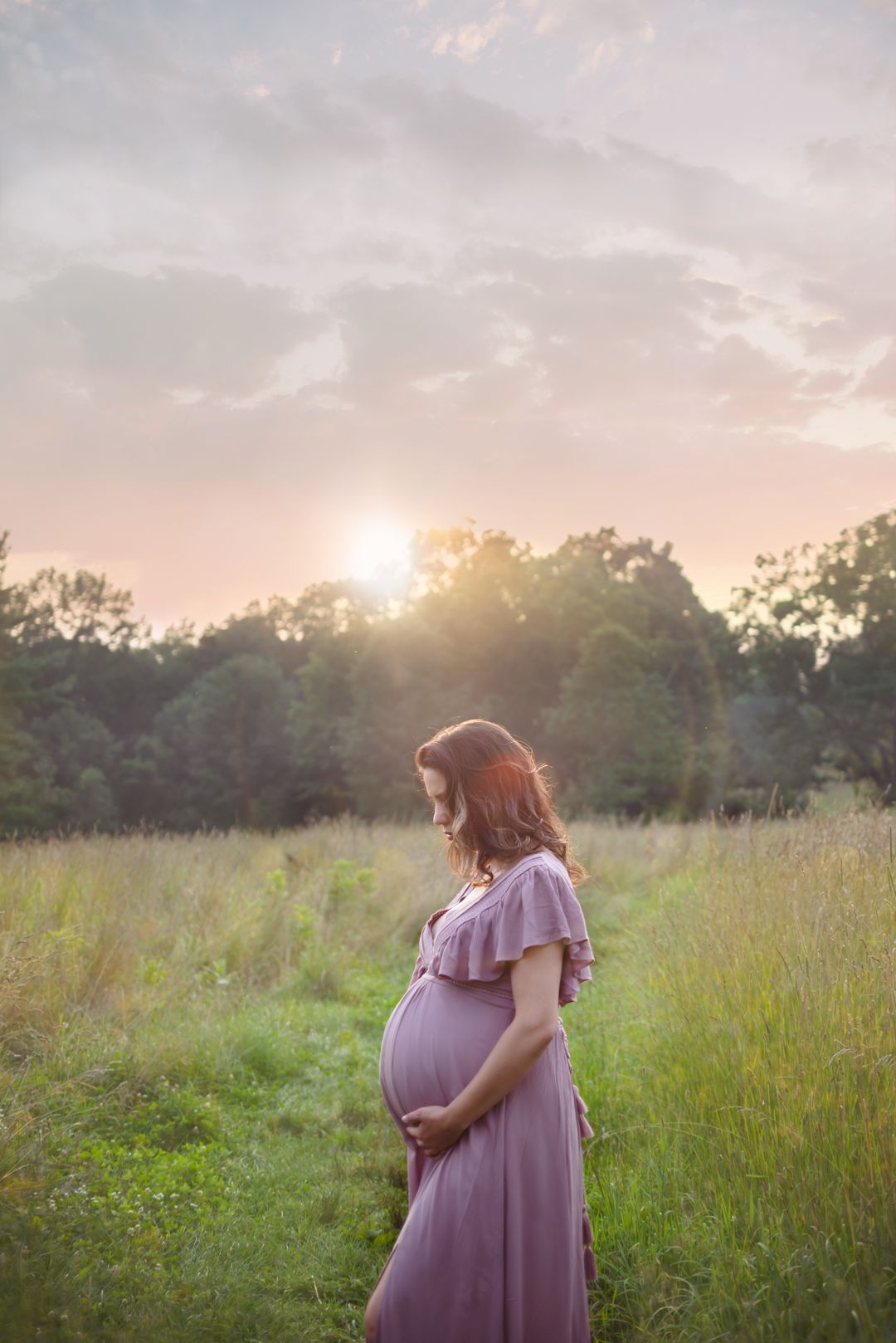 Pregnant woman in flowing purple dress poses in golden field during sunset.