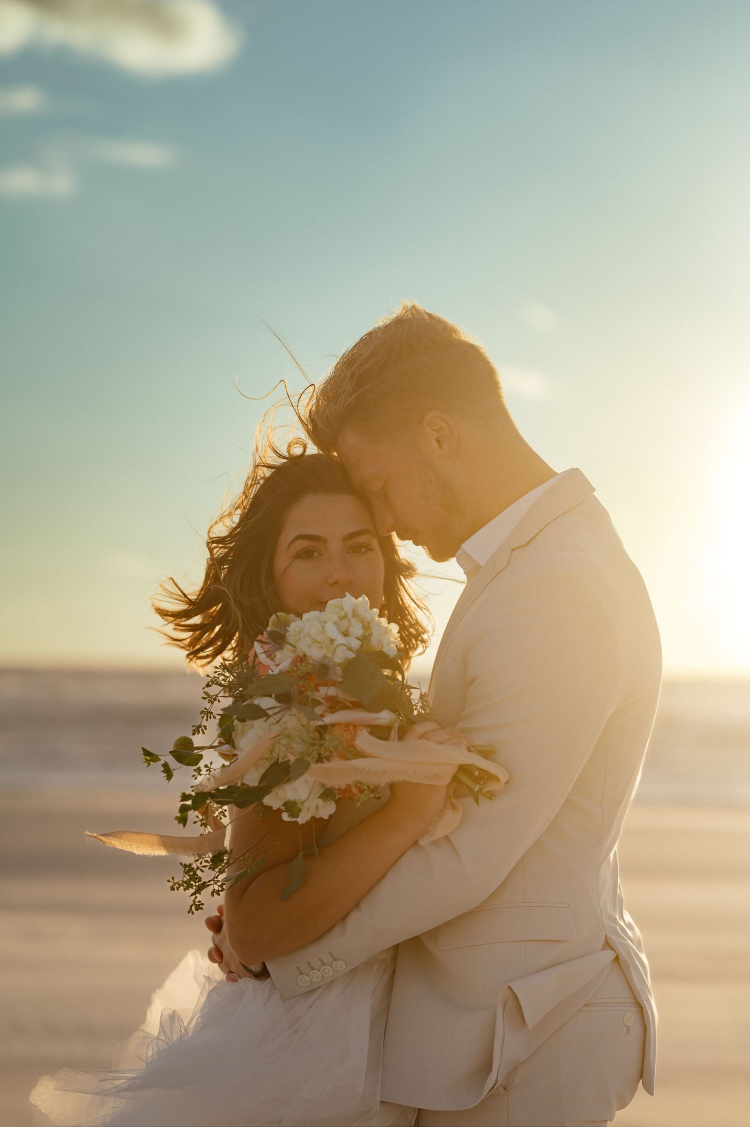 Newlyweds embracing on beach at sunset with bride holding white floral bouquet.