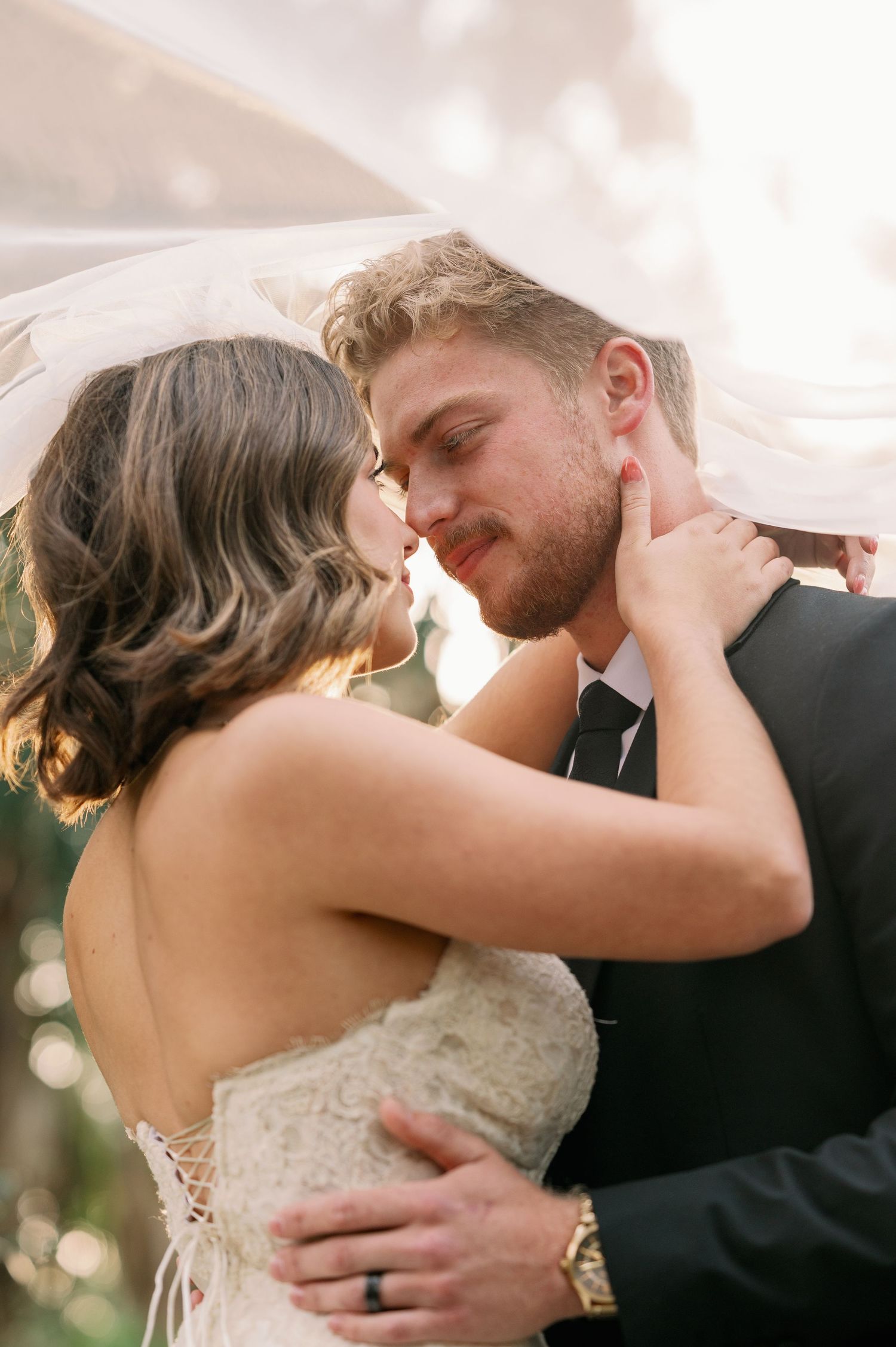 Romantic wedding couple embracing under flowing white veil during outdoor photoshoot.