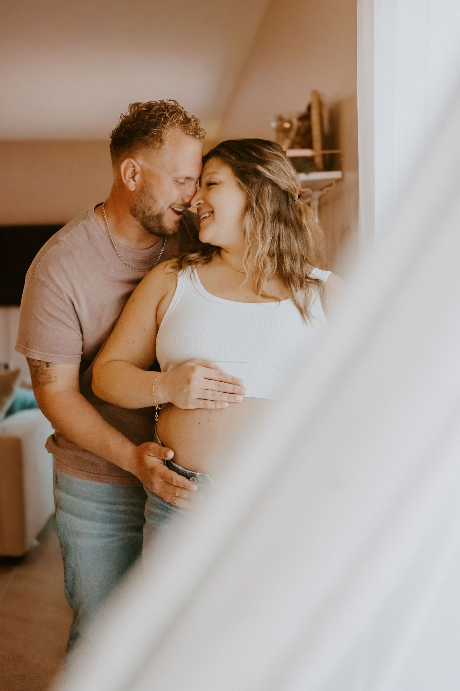 Intimate maternity moment between couple by window in warm lighting.