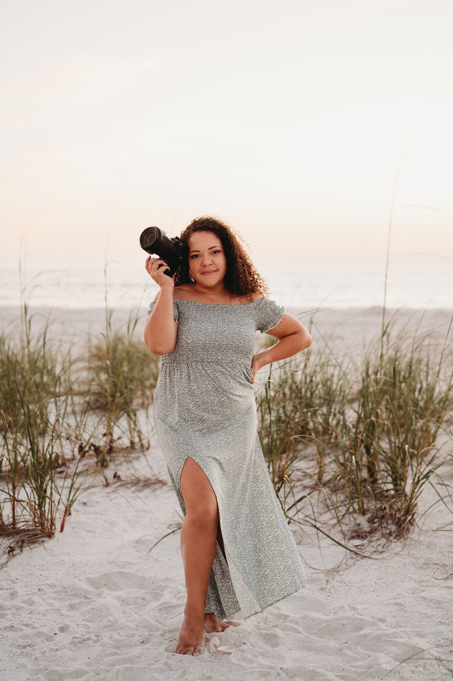 Woman in flowy dress poses with camera on sandy beach during sunset photoshoot.
