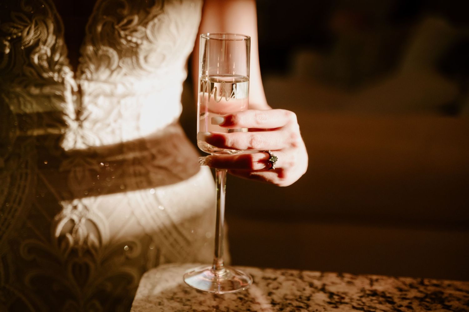 Close-up of wedding dress details with wine glass in warm lighting.