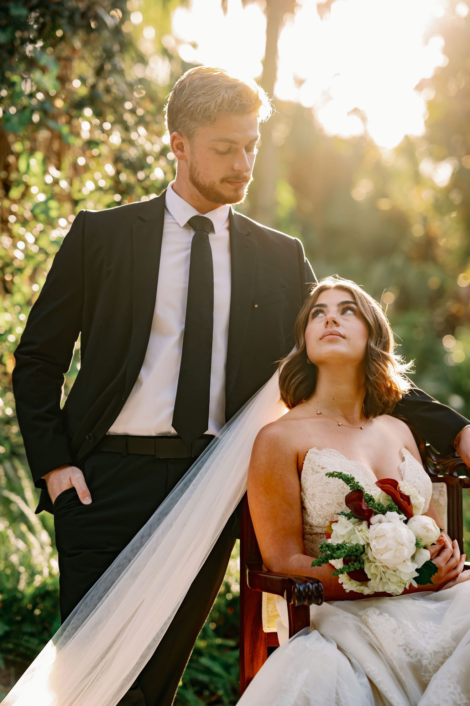 Newlyweds share intimate moments in garden setting with sunlight filtering through trees.