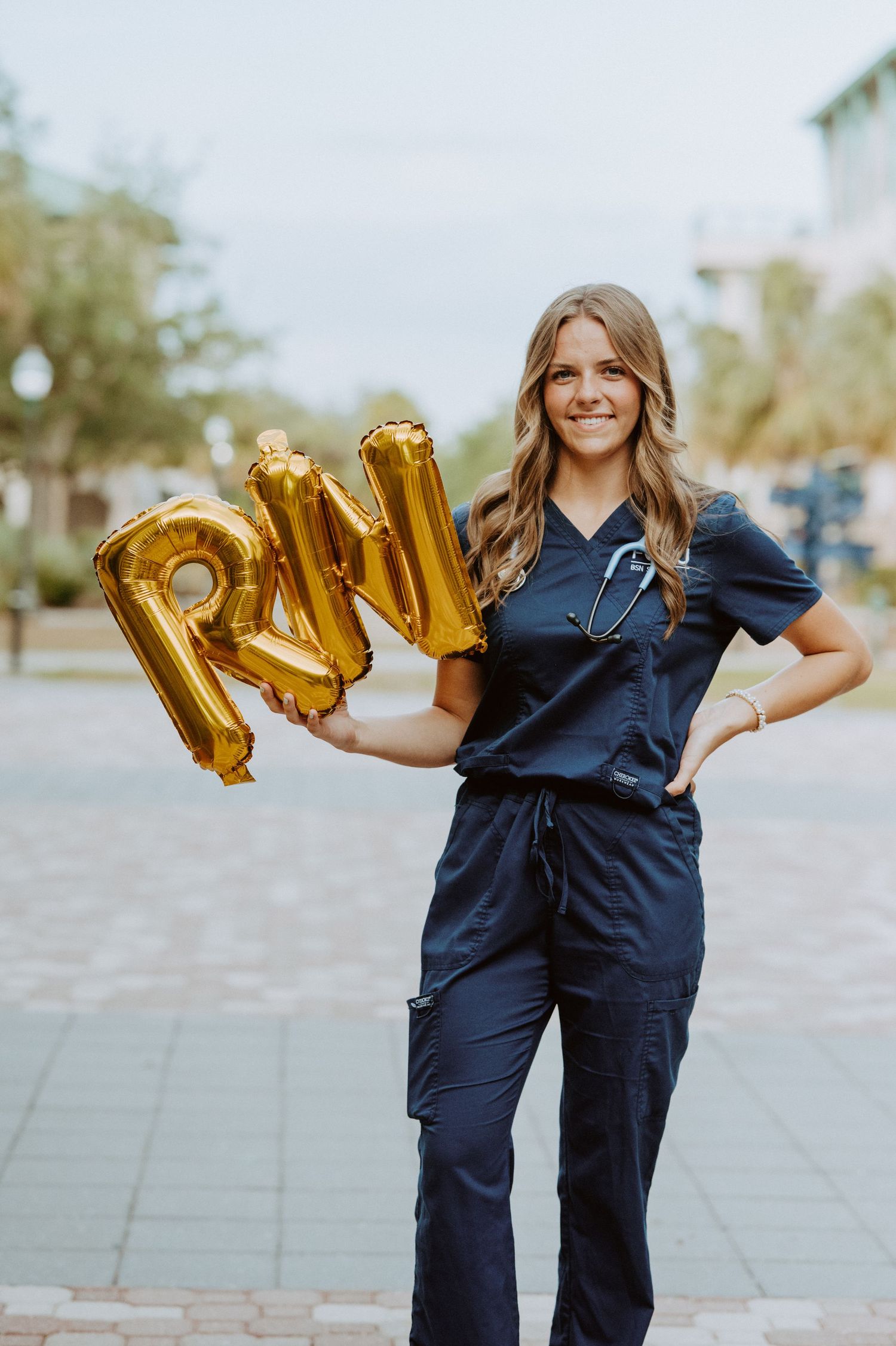 Healthcare worker in navy scrubs poses with gold RN balloon letters in outdoor setting.