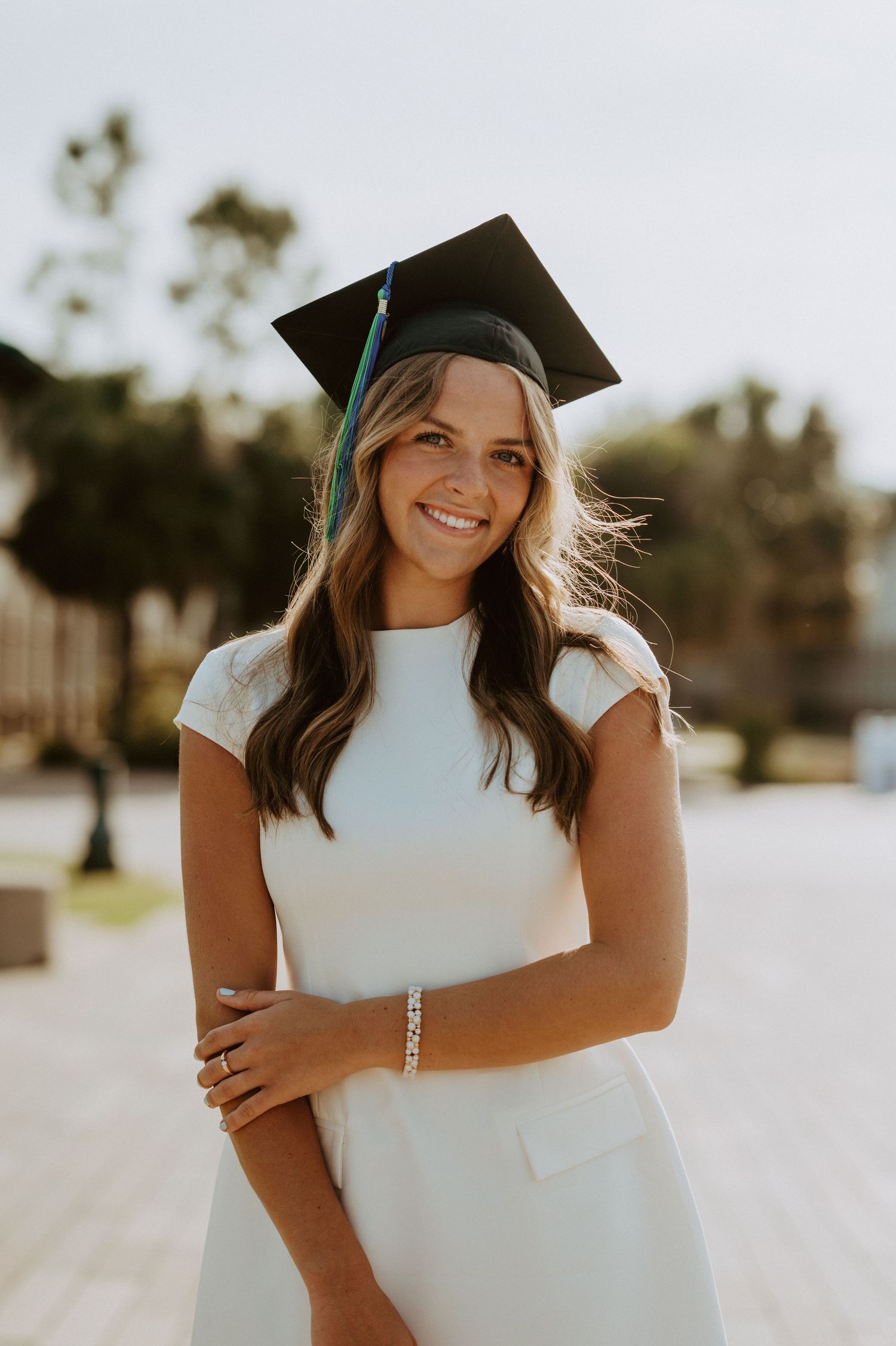 Graduate in white dress and cap poses outdoors in warm sunlight.