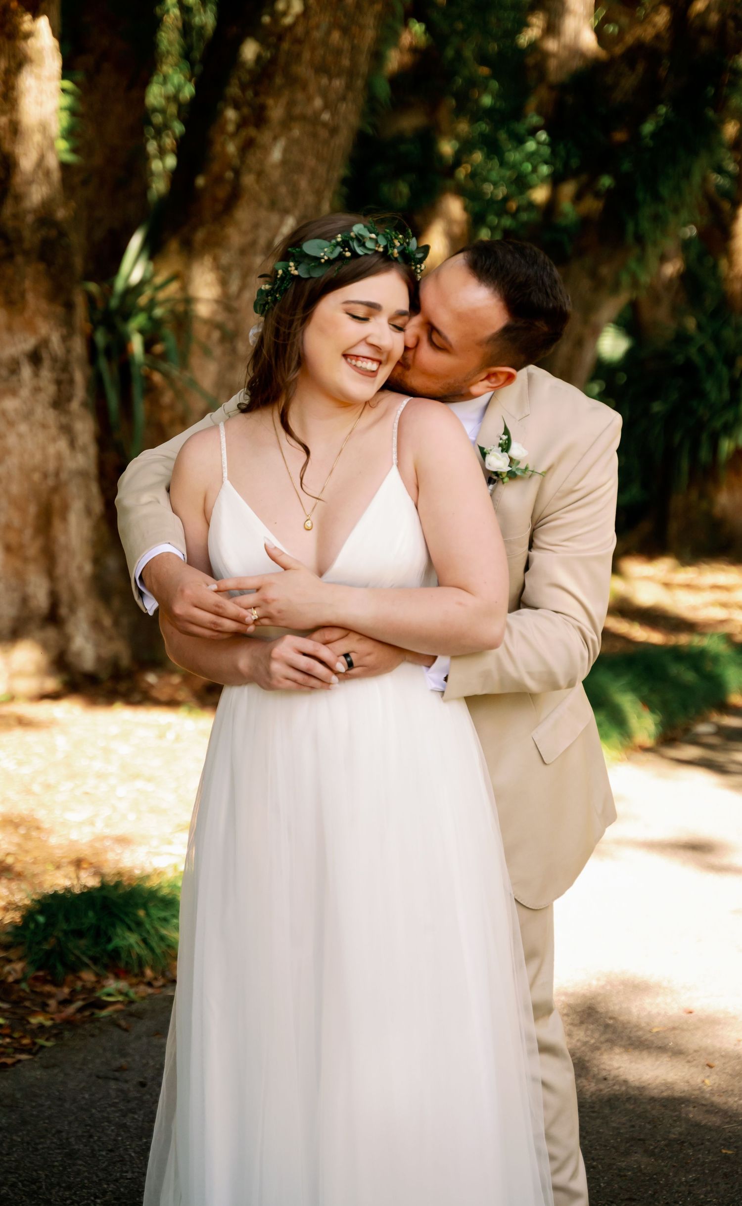 Romantic wedding couple embracing outdoors with natural sunlight filtering through trees.