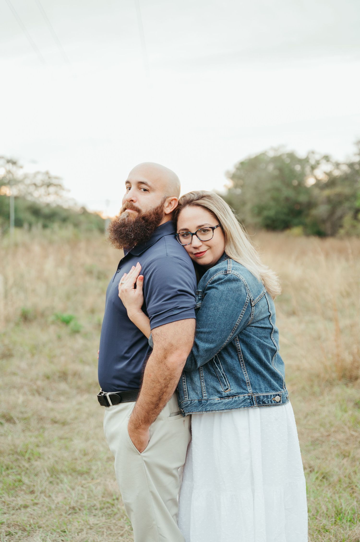 Couple embraces tenderly in a sunlit field wearing denim and casual attire.