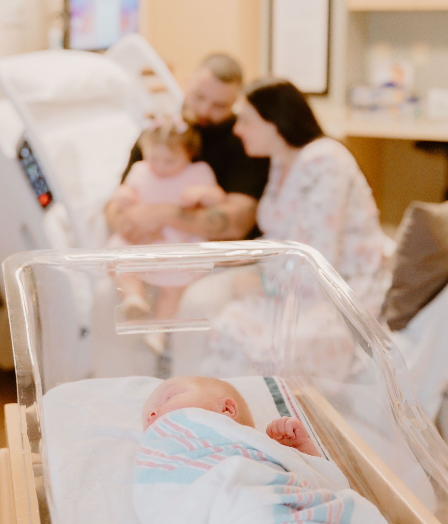 Newborn baby resting in a clear hospital bassinet with medical monitoring equipment.