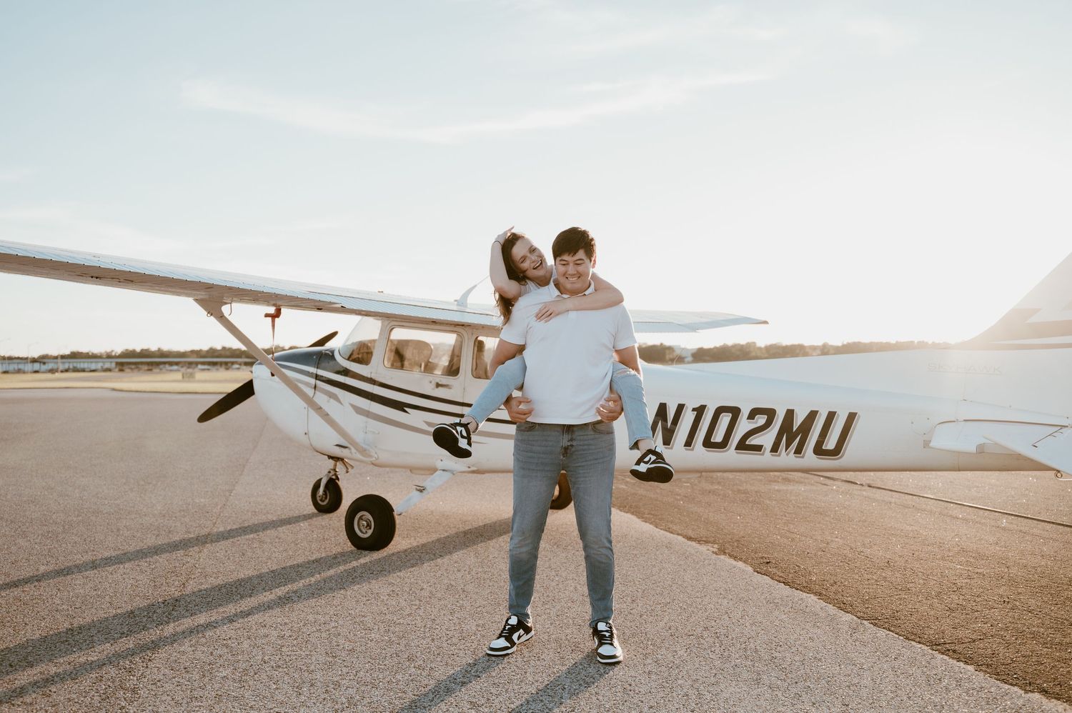 Couple posing playfully in front of a small private airplane on an airfield.
