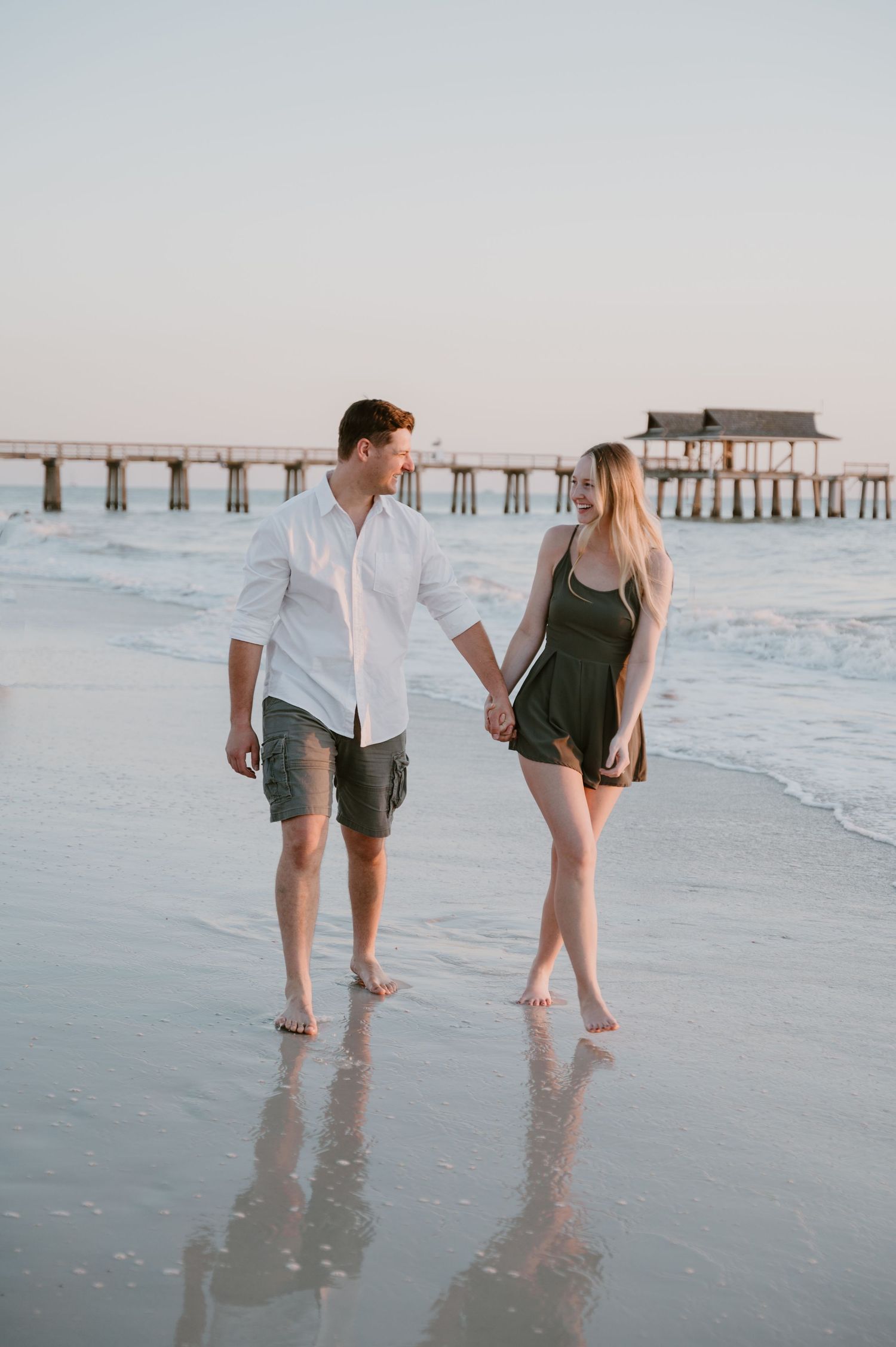 Couple walking hand in hand on beach at sunset with pier in background.