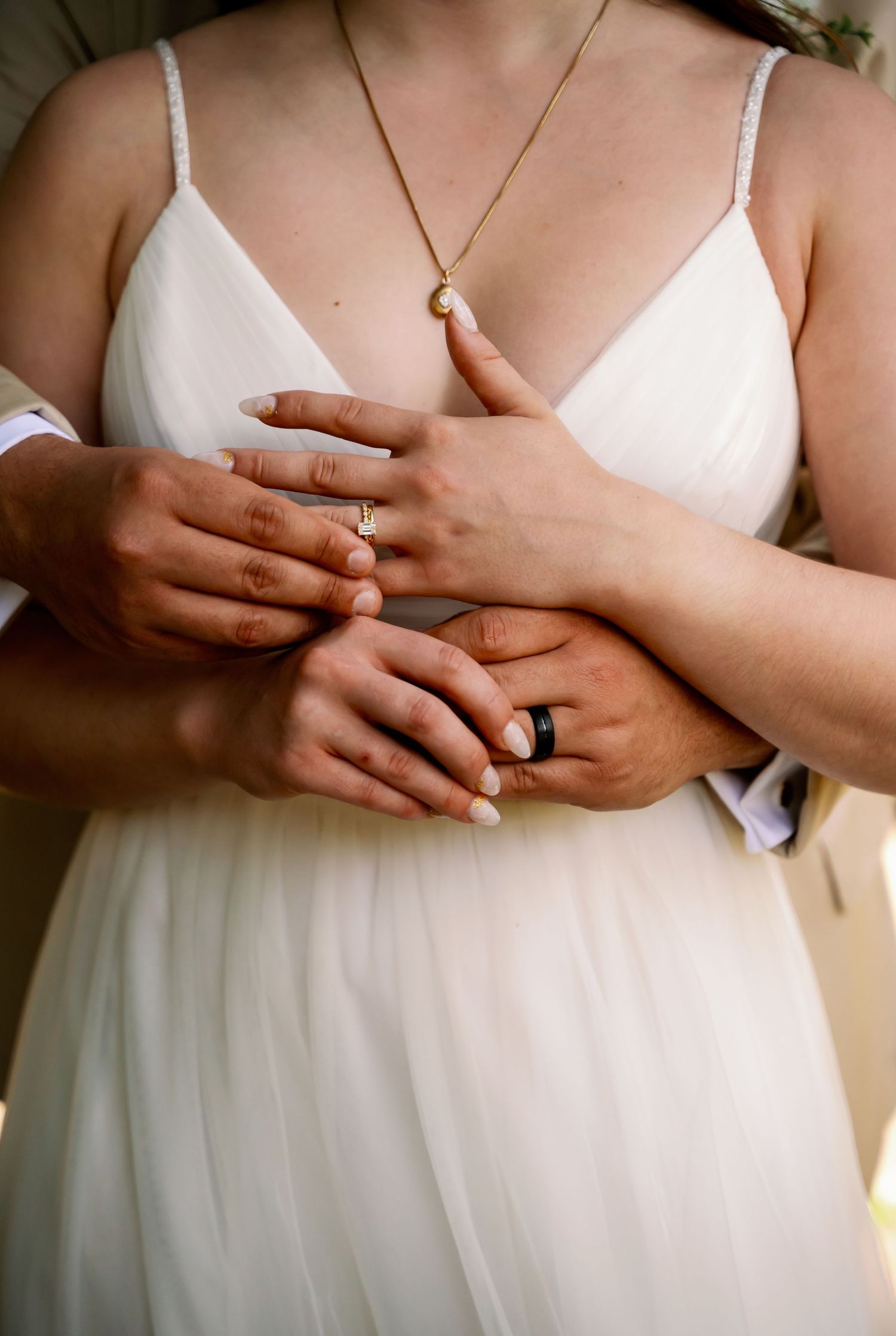 Close-up of hands with wedding rings and crescent moon necklace against white dress.