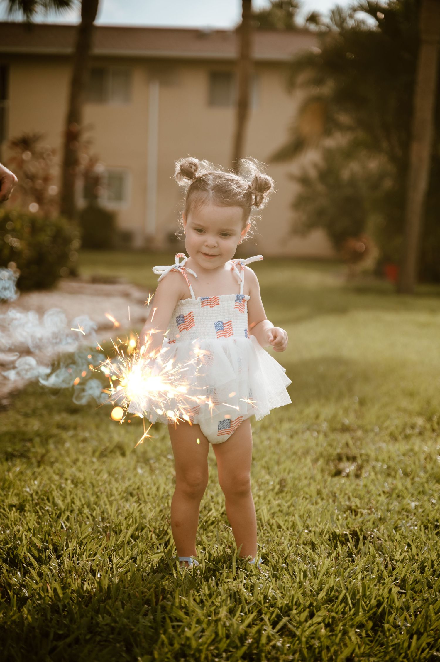 A small child in a white outfit plays with sparklers in the grass during golden hour.