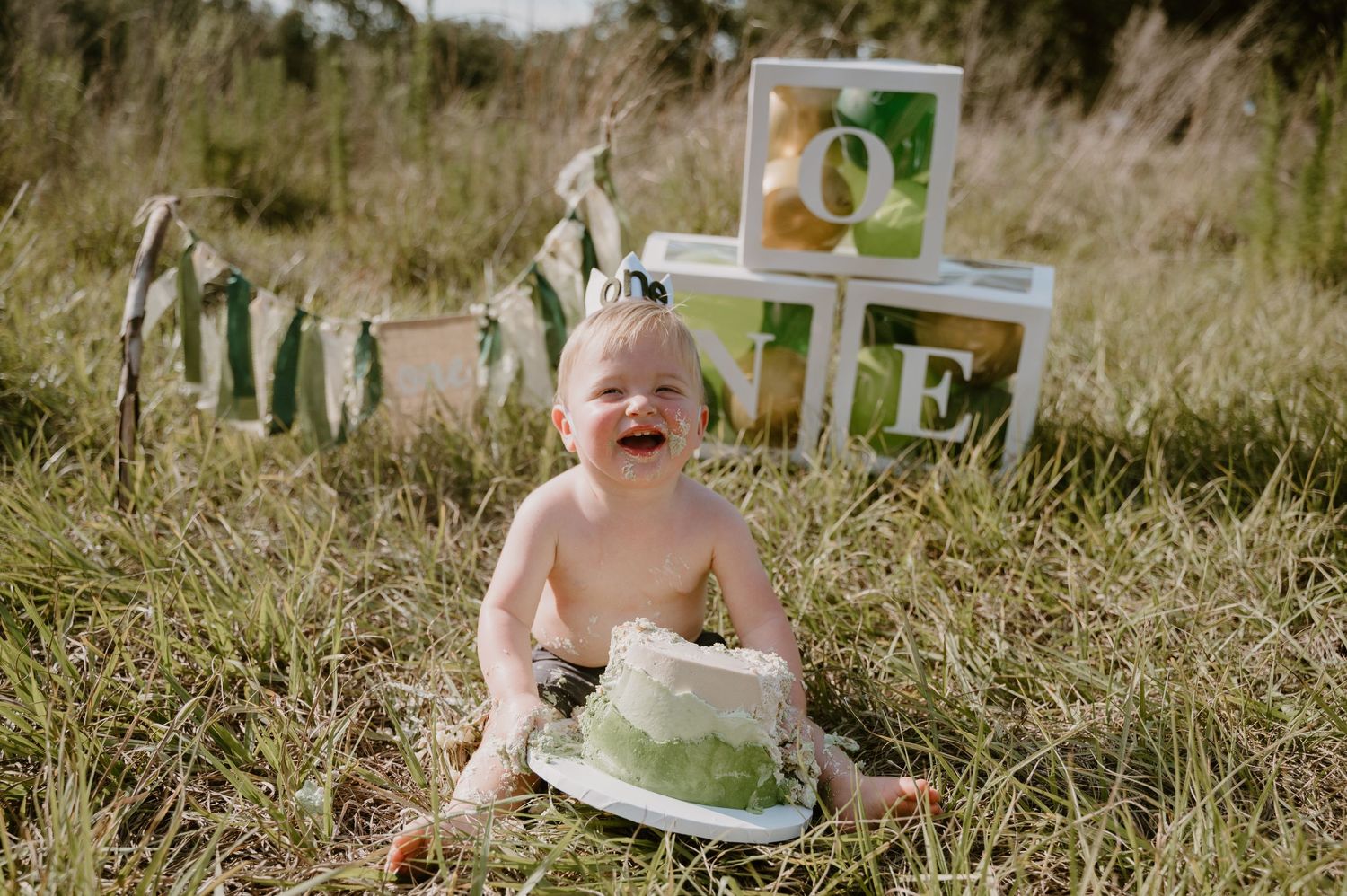 A baby smiles and plays with a green and white cake during what appears to be a first birthday photoshoot.