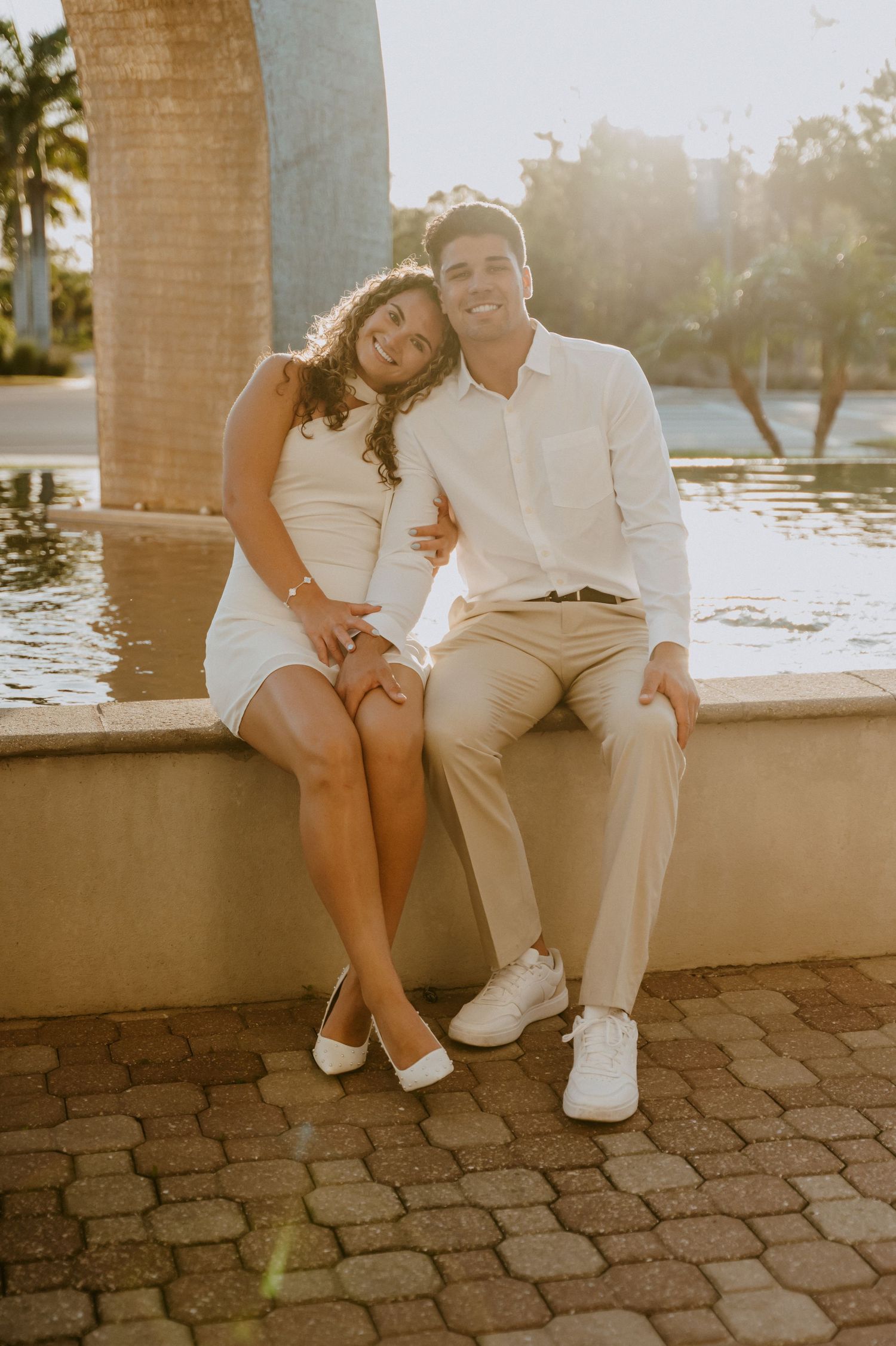 Couple in white clothing sitting together on stone wall at sunset.