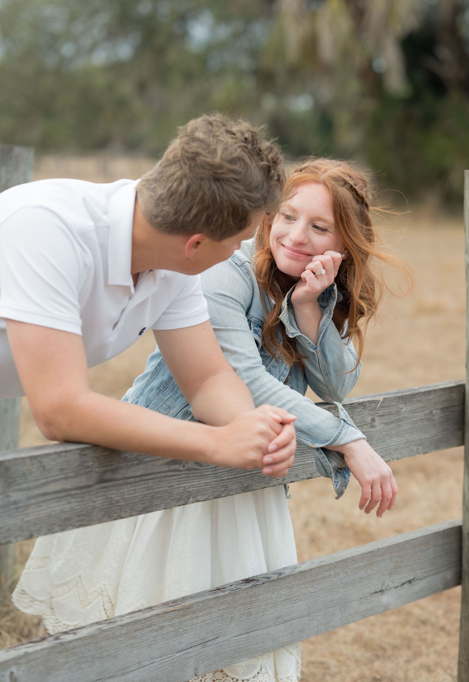 Couple sharing playful moment leaning on rustic wooden fence in countryside setting.