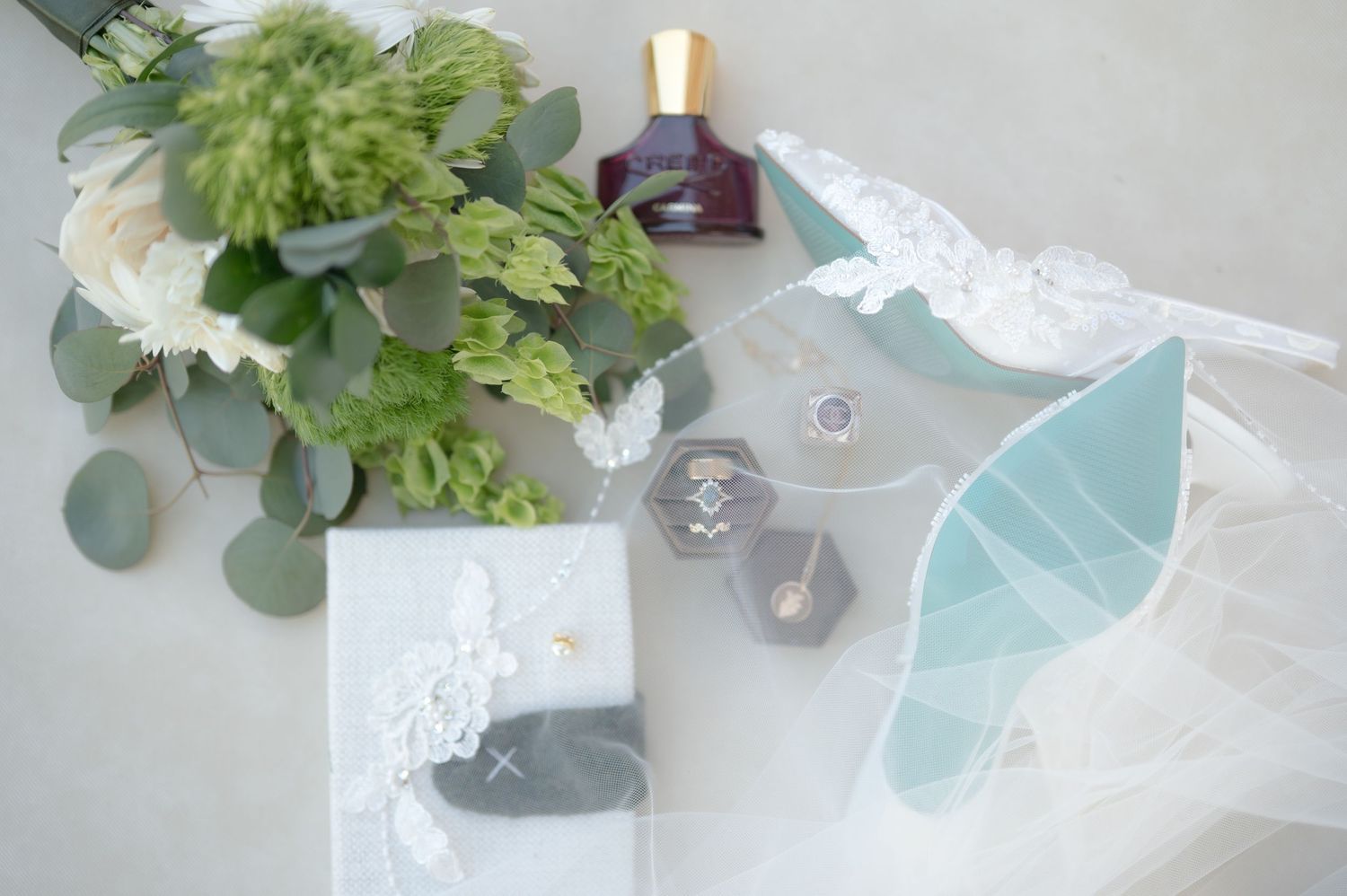 Elegant wedding flat lay with green floral bouquet, ring boxes, perfume bottle, and lace veil on gray background.