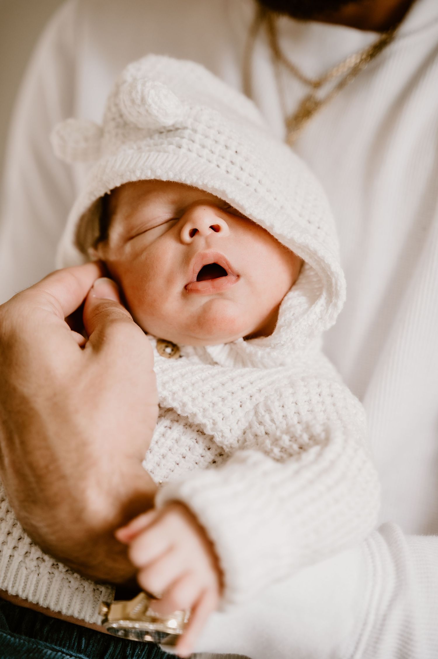 Sleeping baby wearing white knit hat and sweater being held gently.