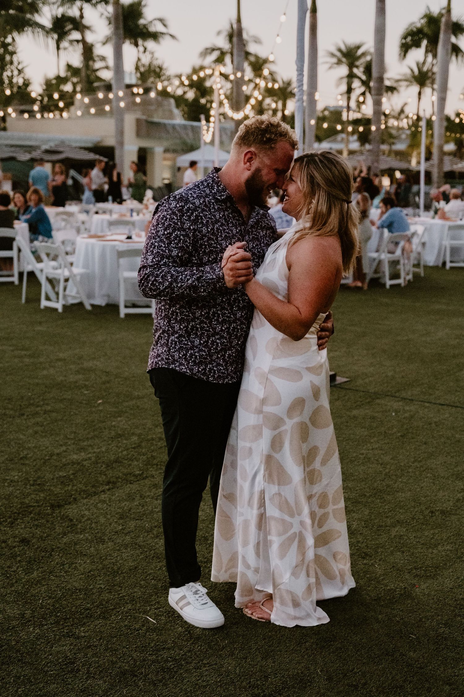 Couple dancing at outdoor reception with string lights and palm trees in background.