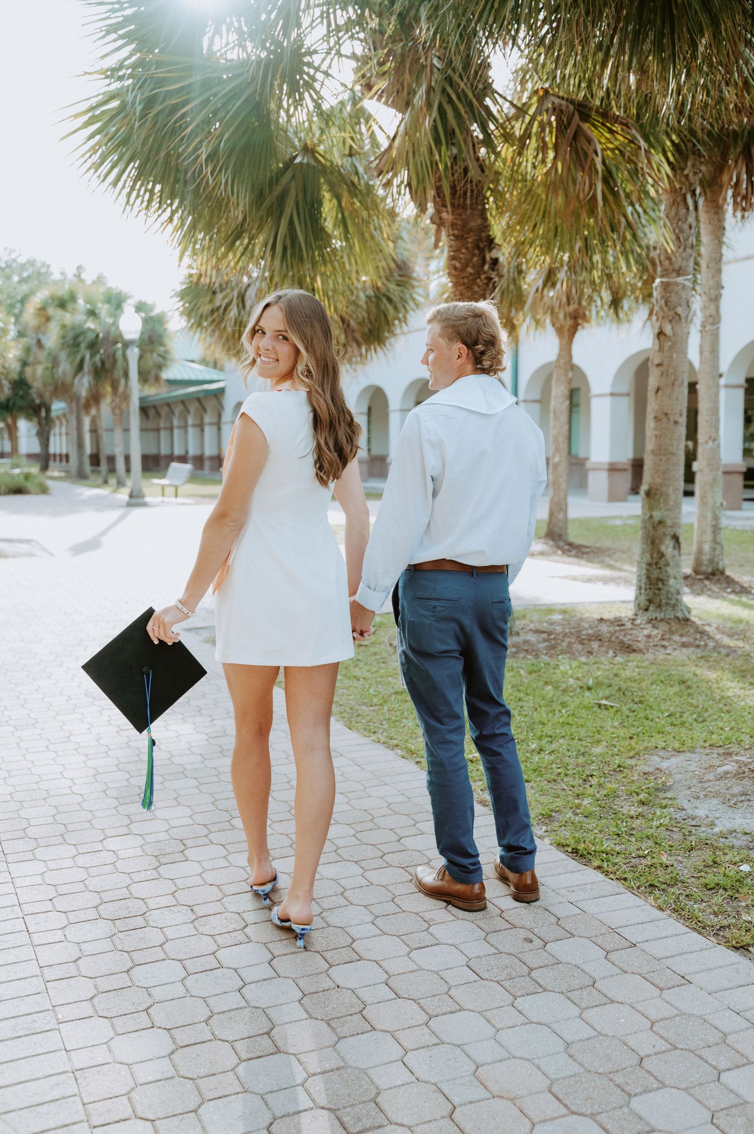 A graduate walks with companion along a palm-lined path holding diploma.