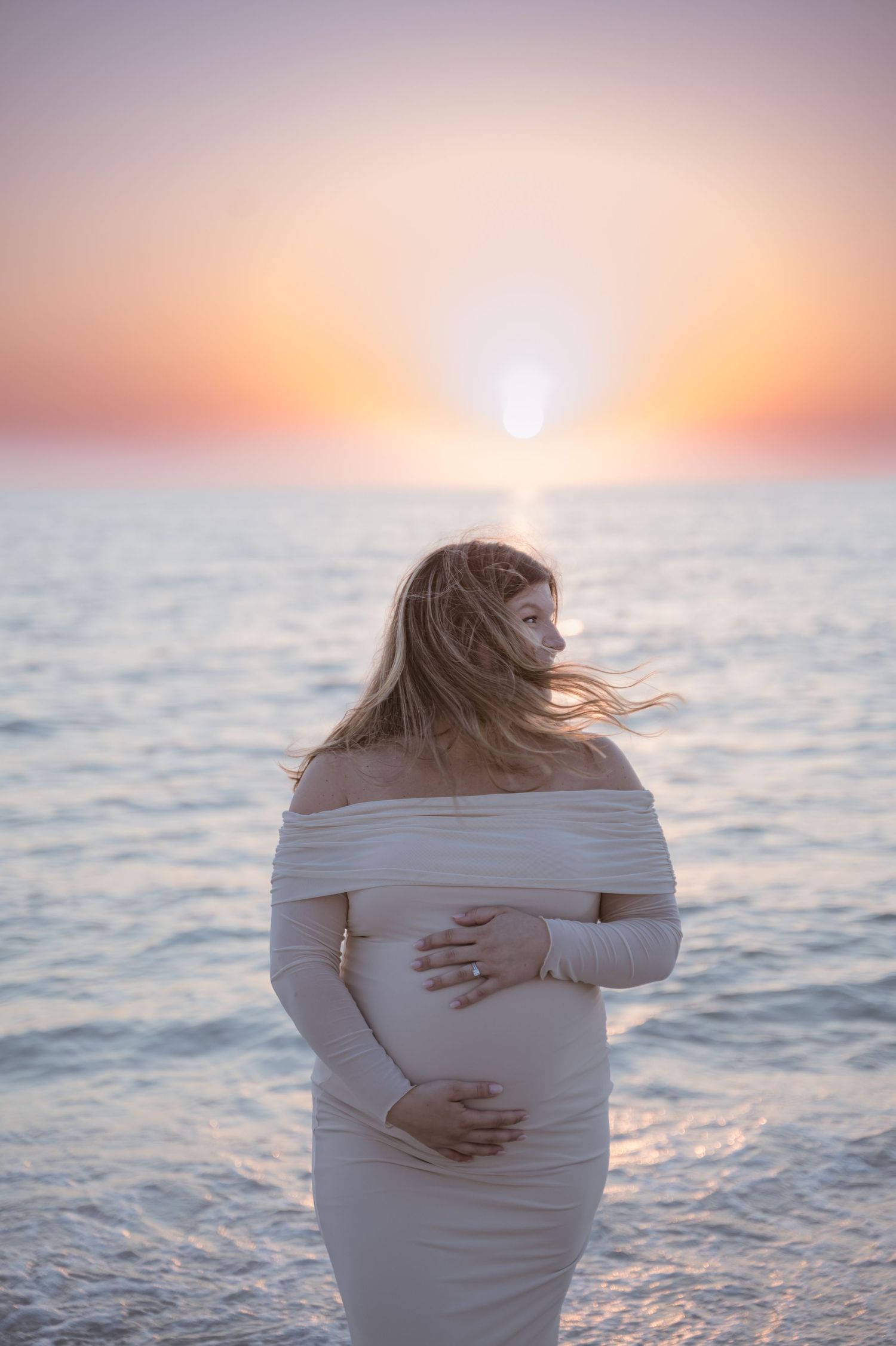 Silhouette of pregnant woman in white dress against ocean sunset.