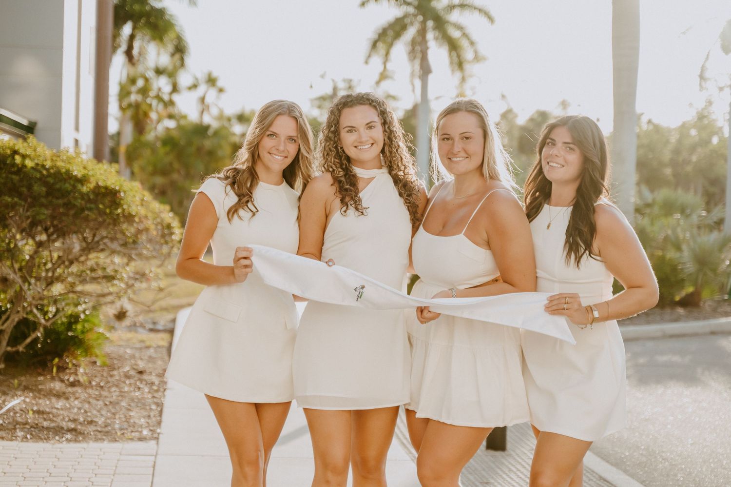 Four friends in matching white dresses pose together on a sunny coastal path.