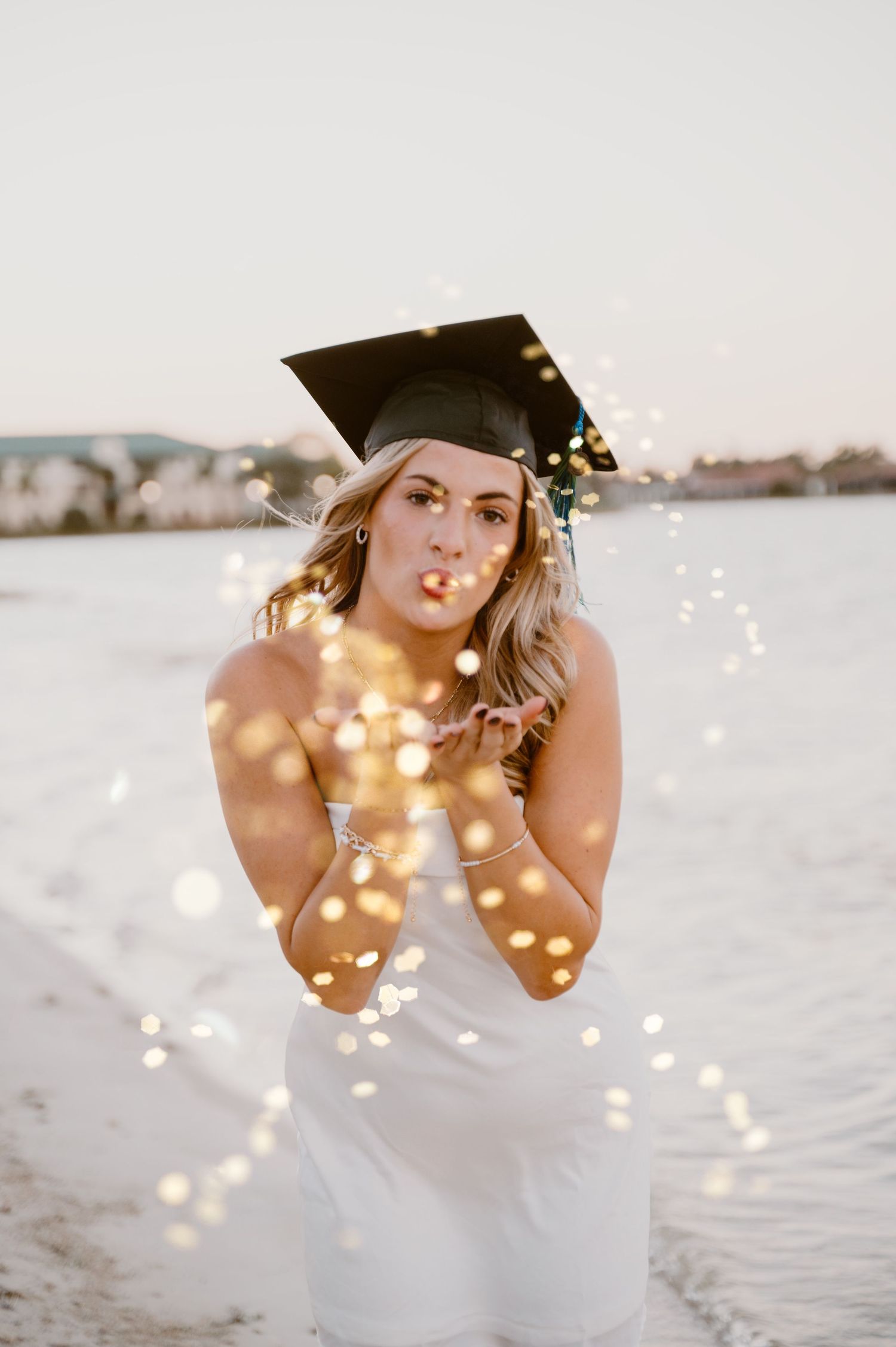Graduate in white dress poses with sparklers on beach during sunset celebration photoshoot.